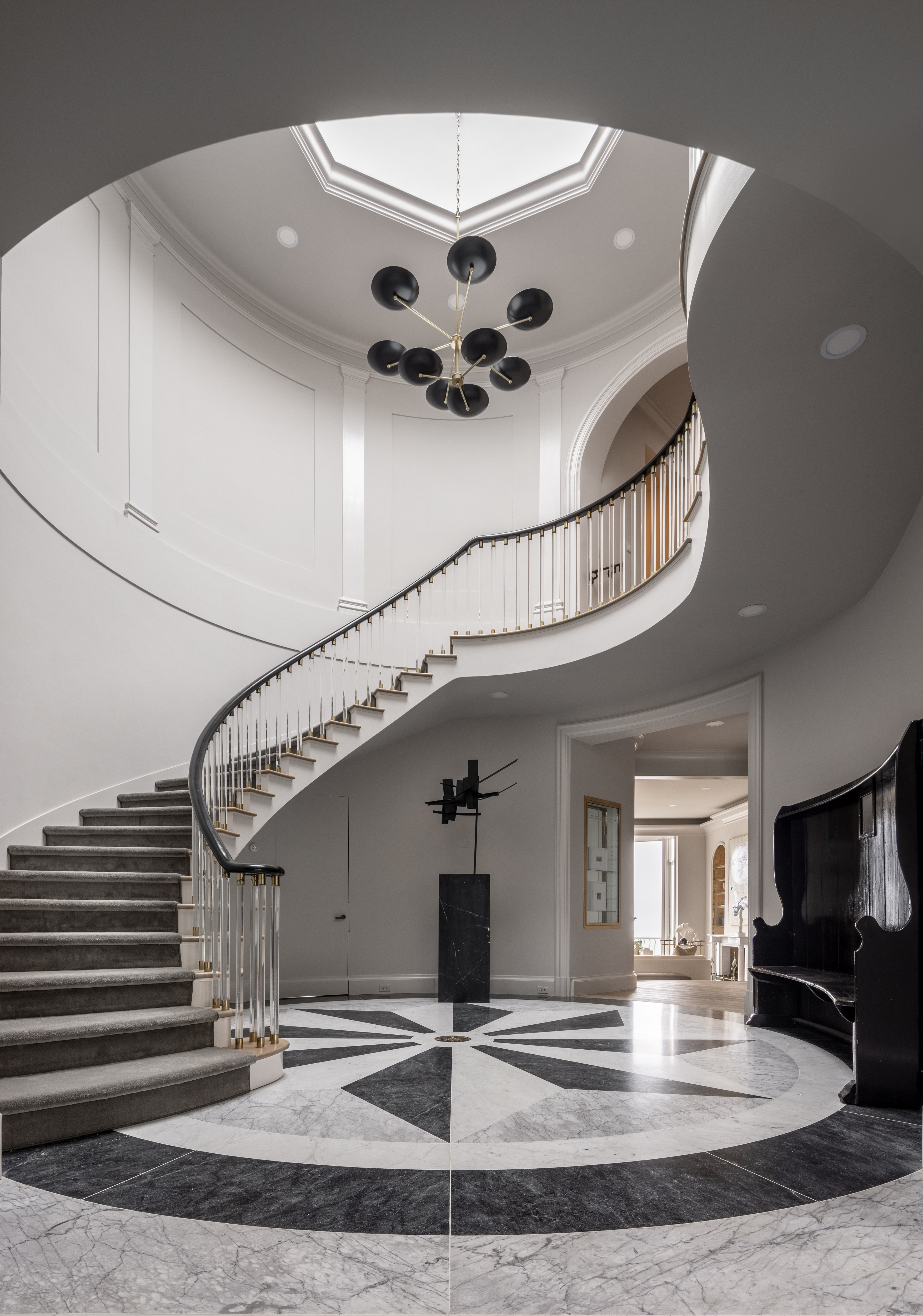 The image shows a grand, modern foyer with a curved staircase, black-and-white geometric marble floor, circular chandelier, and minimalist décor in neutral tones.