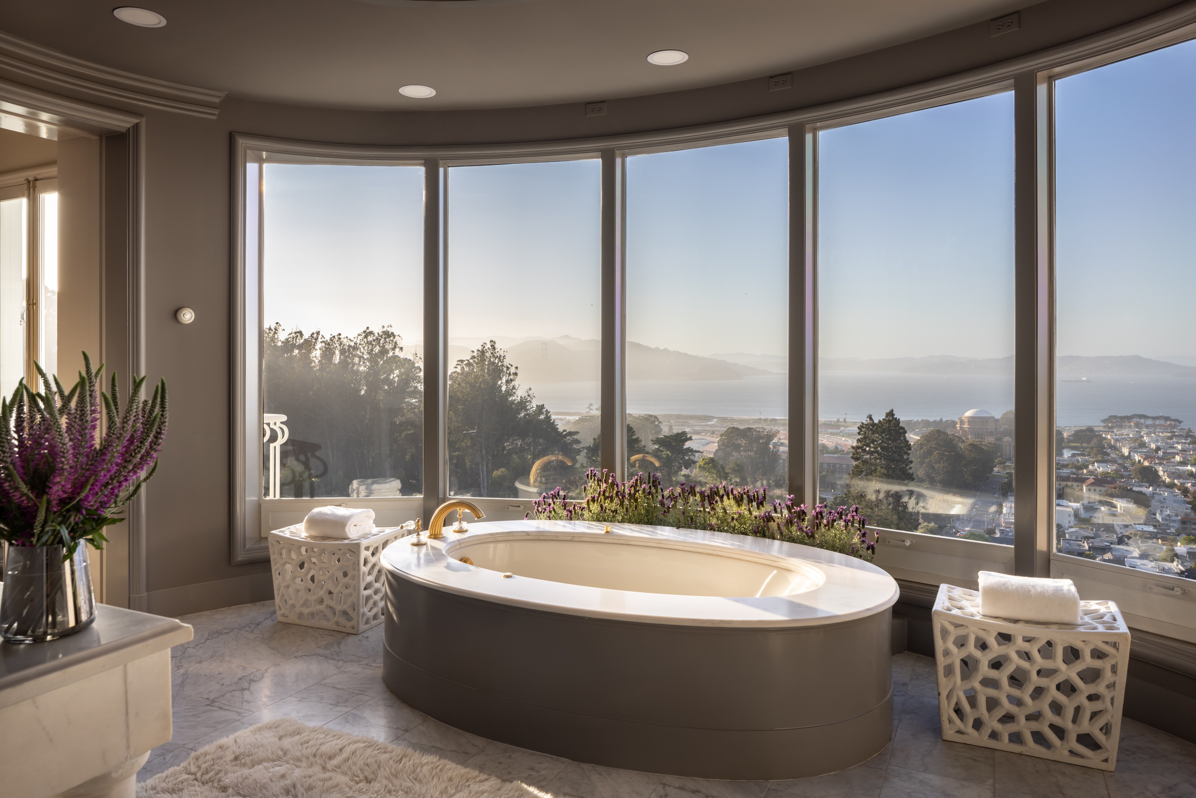 A luxurious bathroom features a large oval tub with a golden faucet and surrounding flowers, flanked by white geometric side tables with towels, set against wide windows showcasing a scenic ocean view.