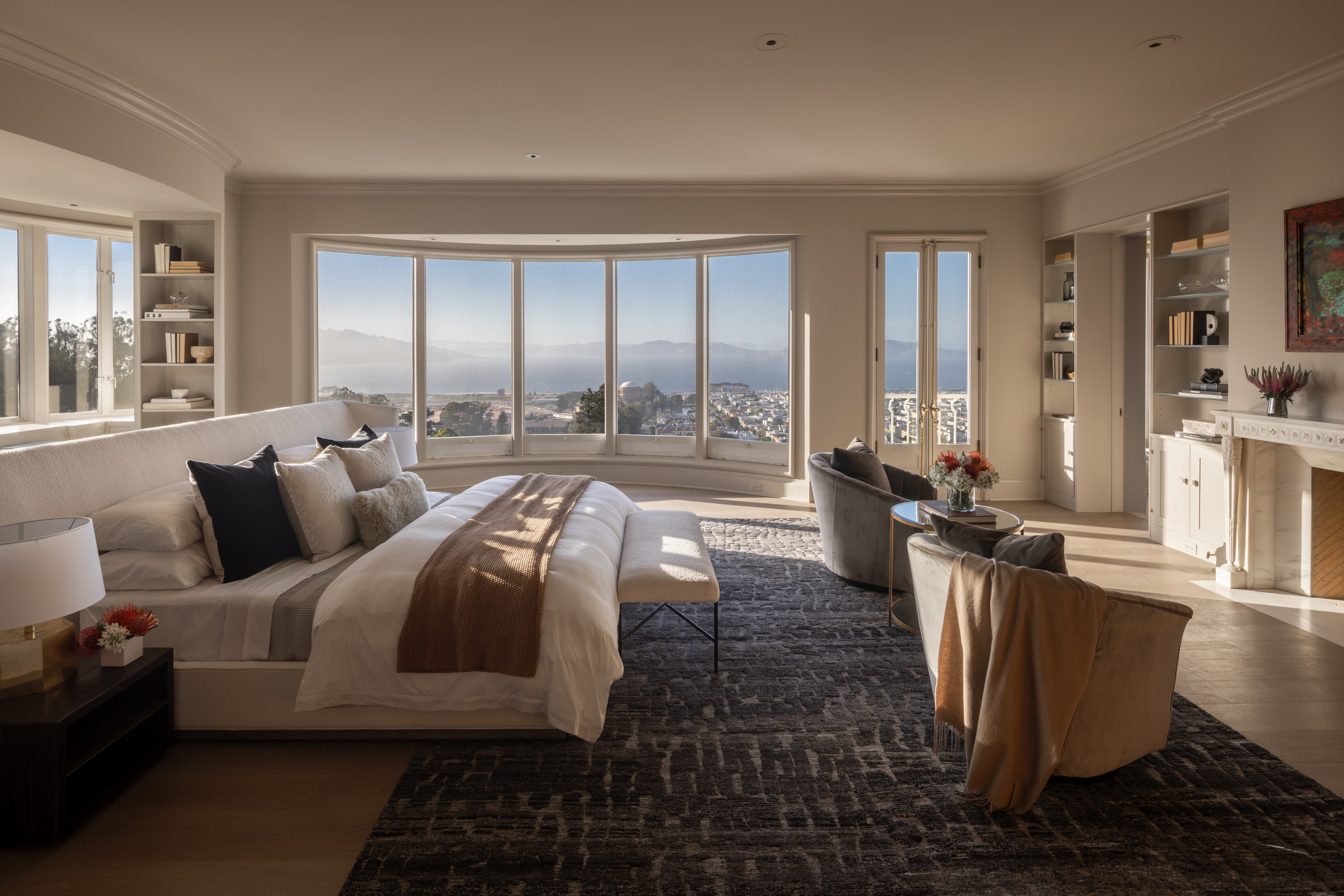 A spacious bedroom features a large bed facing wide windows with a scenic city and water view. It includes cozy seating, a fireplace, built-in shelves, and elegant decor.