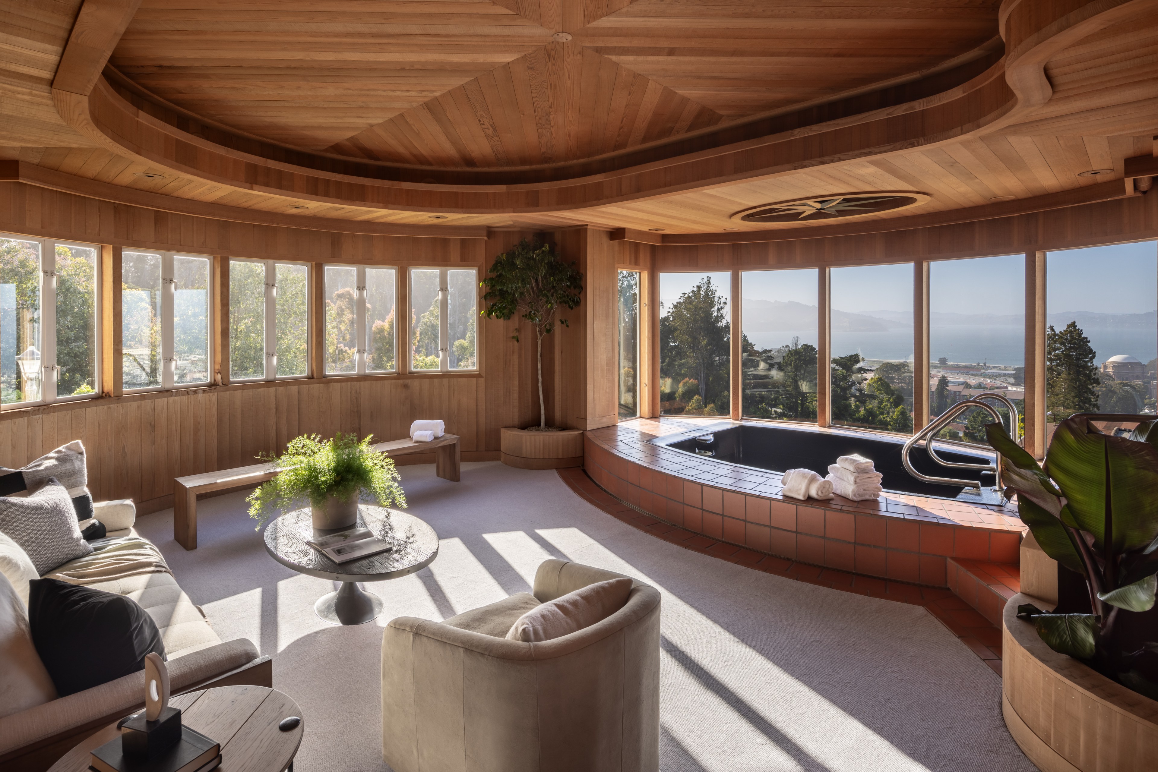 The room features a wooden interior with large windows overlooking a scenic view. It has a cozy seating area with a round table, plush chairs, and a built-in hot tub.