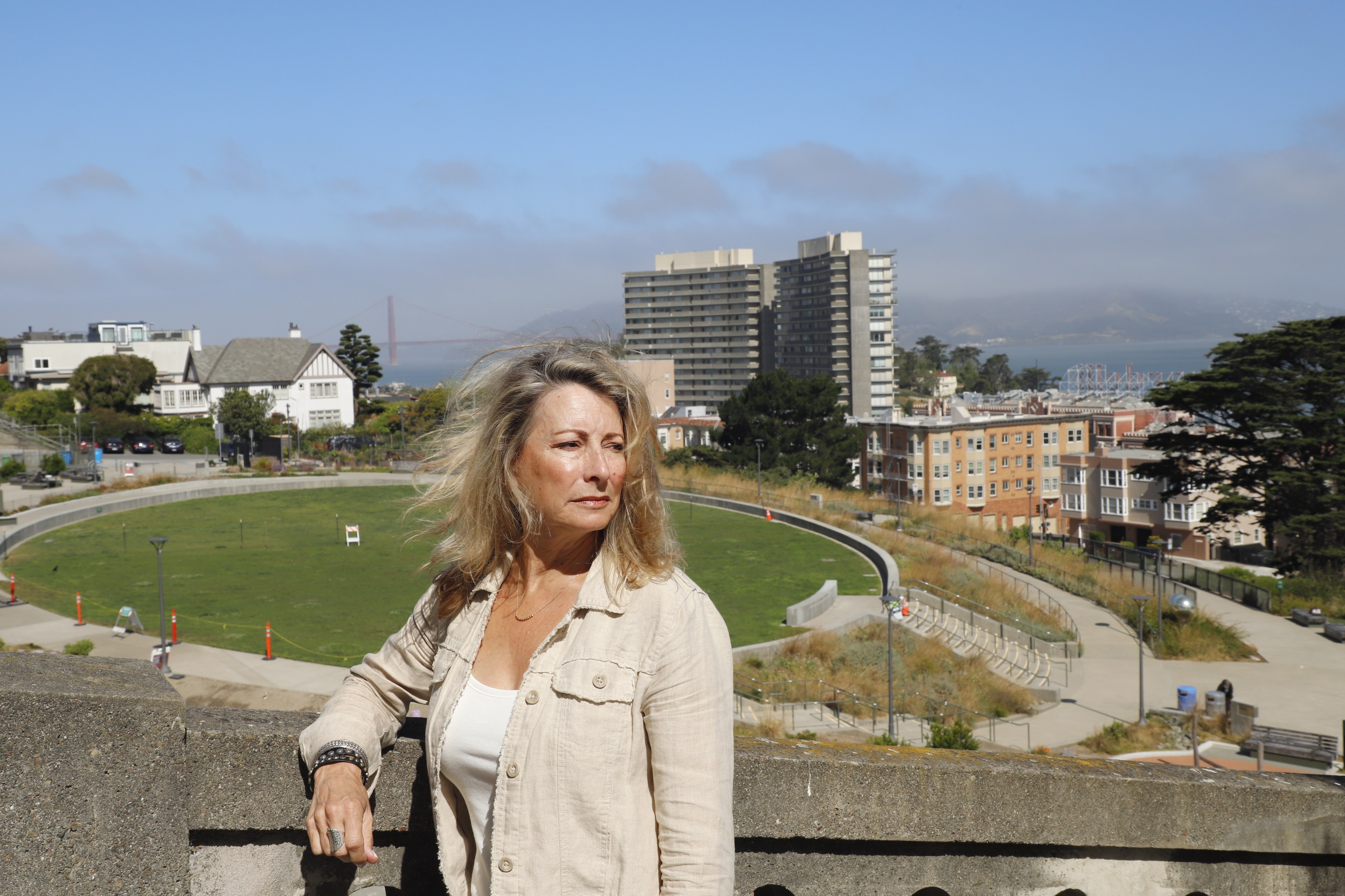 A woman with long hair stands near a concrete railing, overlooking a park and cityscape with a mix of buildings, including tall apartments and houses. The Golden Gate Bridge is visible in the background.