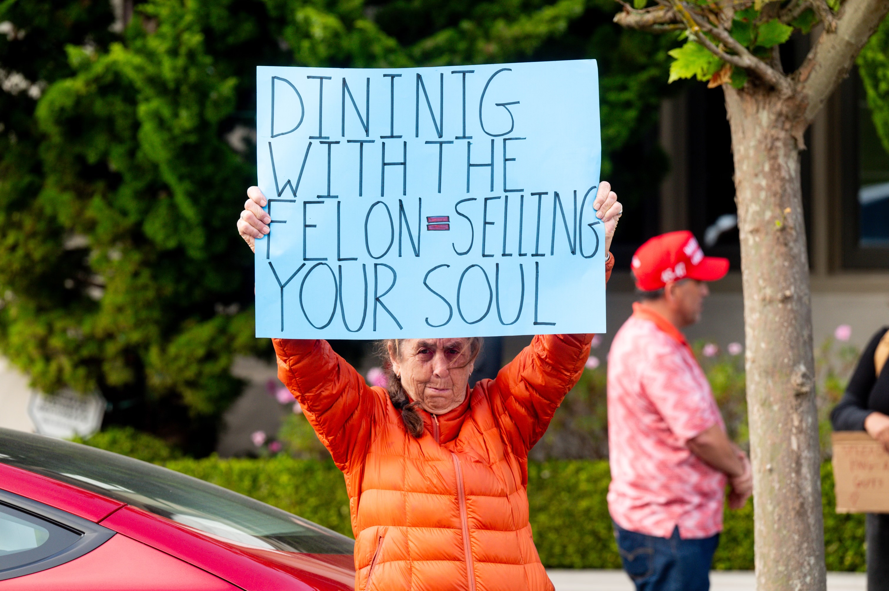 A person in an orange jacket holds a sign saying &quot;DINING WITH THE FELON = SELLING YOUR SOUL&quot; while standing outside, with others in the background.