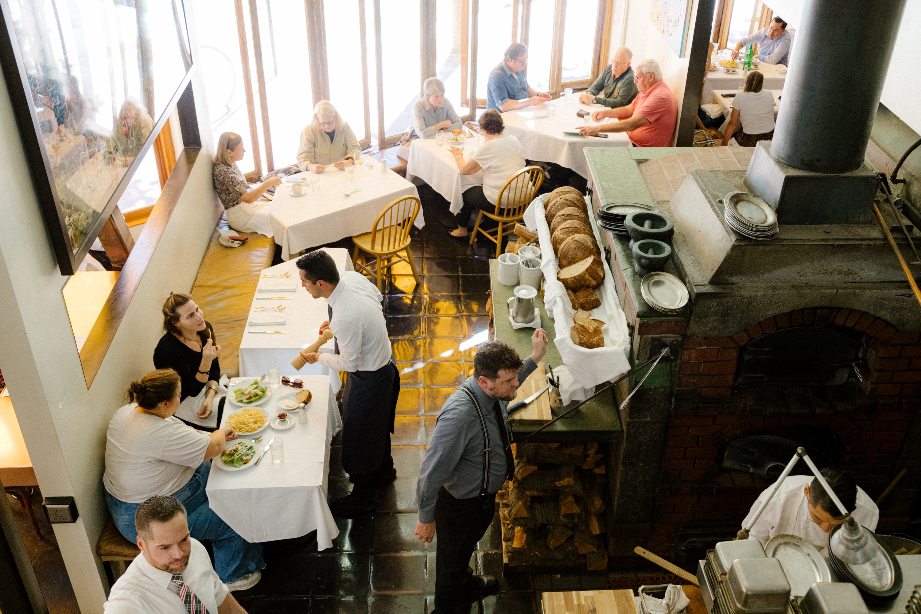 People dine at white-clothed tables in a cozy restaurant with large windows. Bread and dishes fill a counter by a brick oven, where staff serve and prepare food.