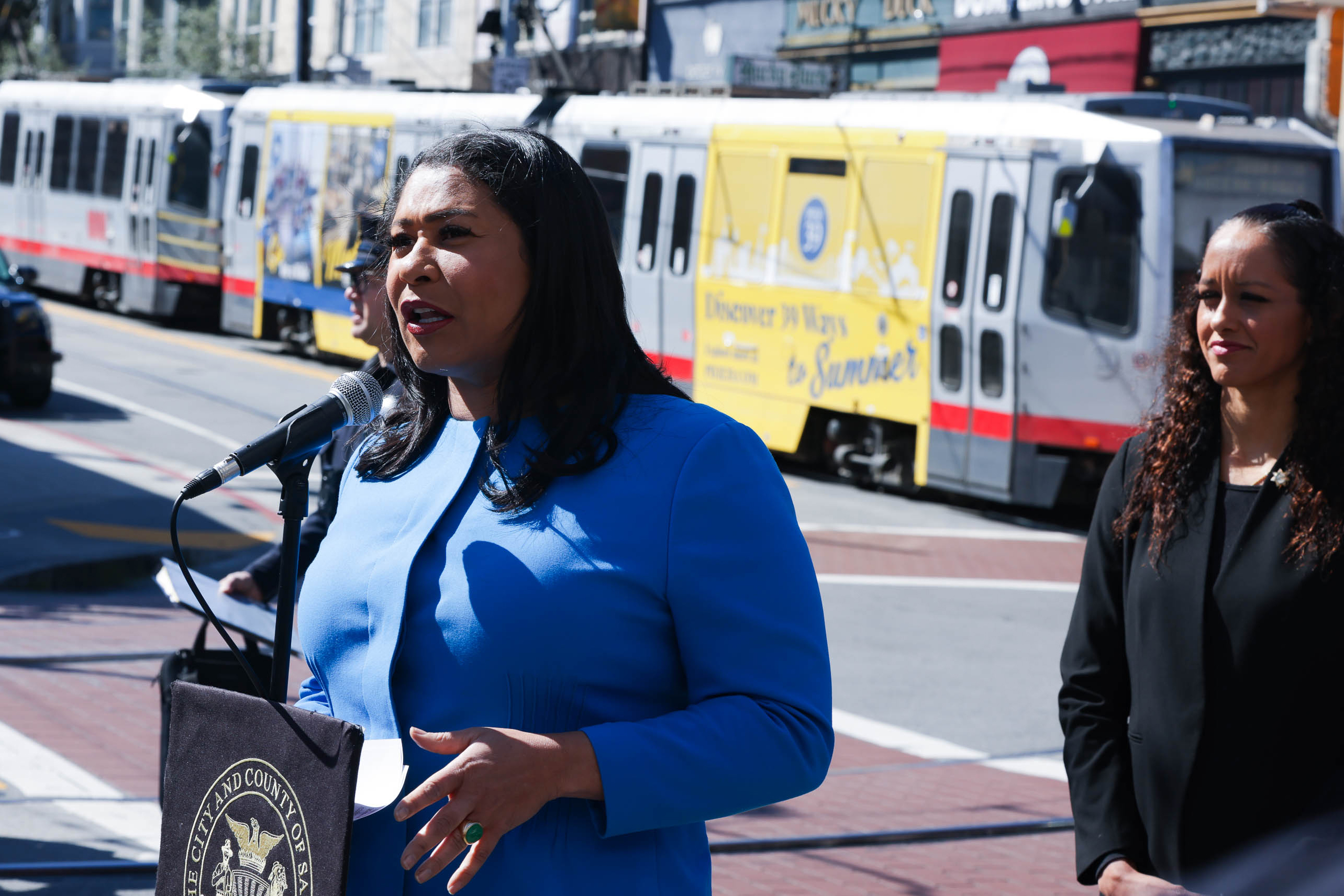 A woman in a blue suit speaks into a microphone at an outdoor event, while another woman in black stands beside her. A city light rail train is in the background.