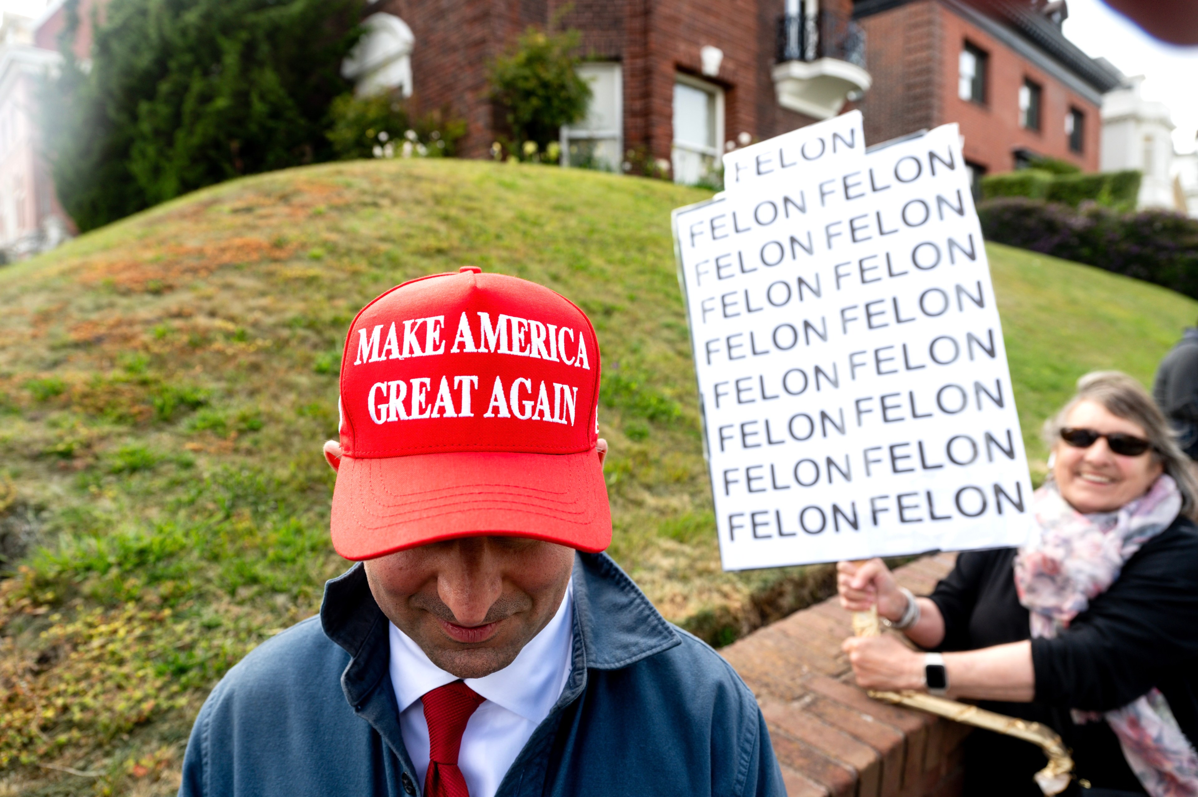 A person wearing a red &quot;Make America Great Again&quot; hat is looking down, while behind them, another person holds a sign repeatedly saying &quot;FELON.&quot;