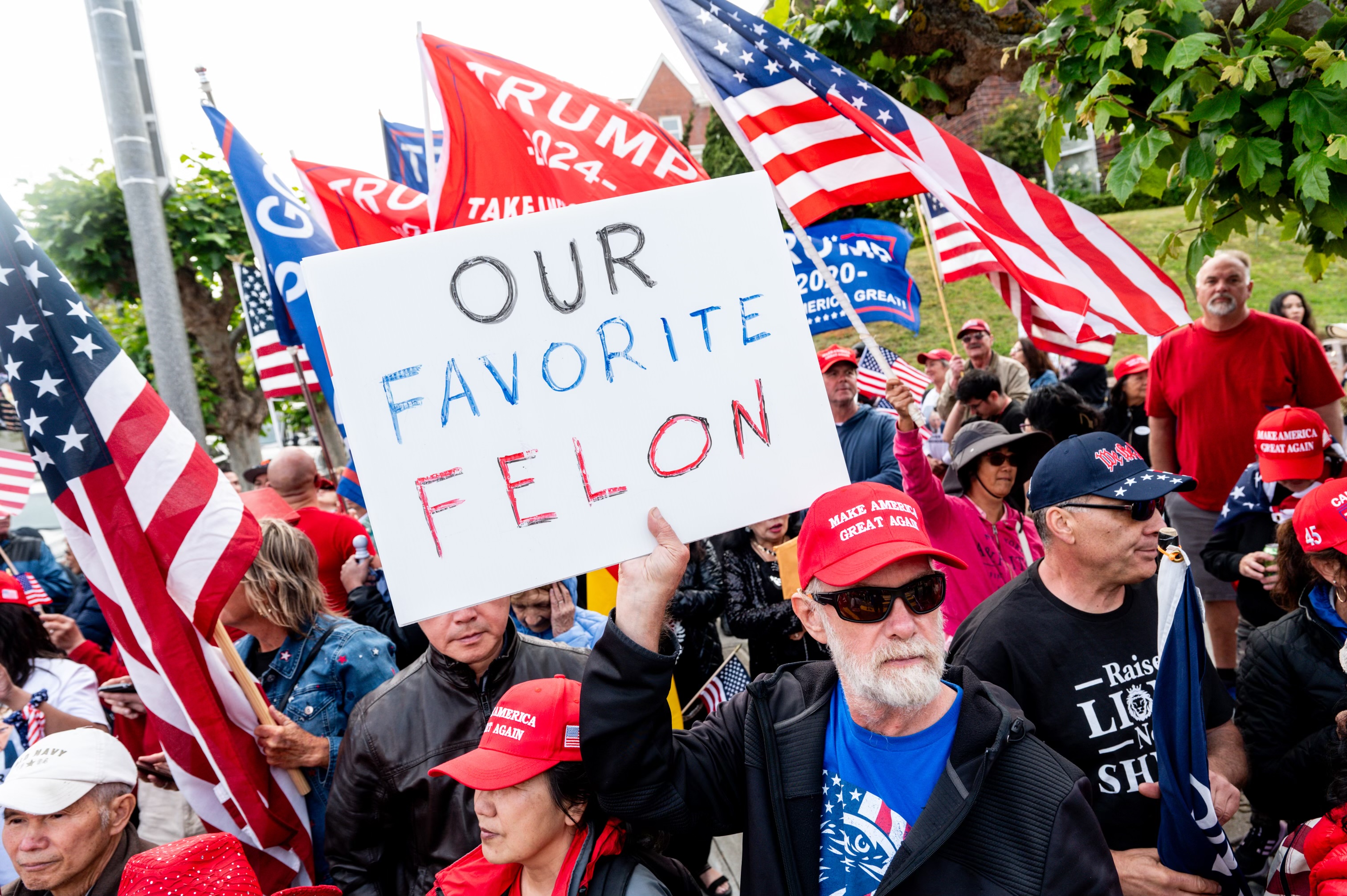 A crowd of people, many wearing red "Make America Great Again" hats, hold American flags and a sign that reads "OUR FAVORITE FELON" at a rally.
