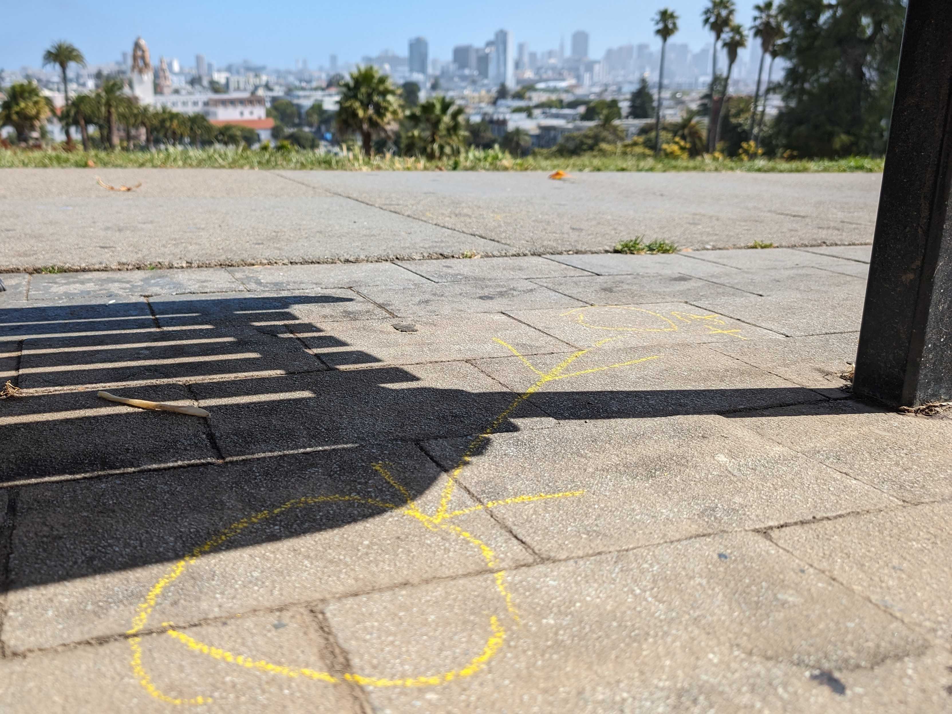 The image shows a city skyline from a park, with palm trees and a shadow on the ground. A yellow chalk drawing on the pavement says &quot;LOVE&quot; with a heart.