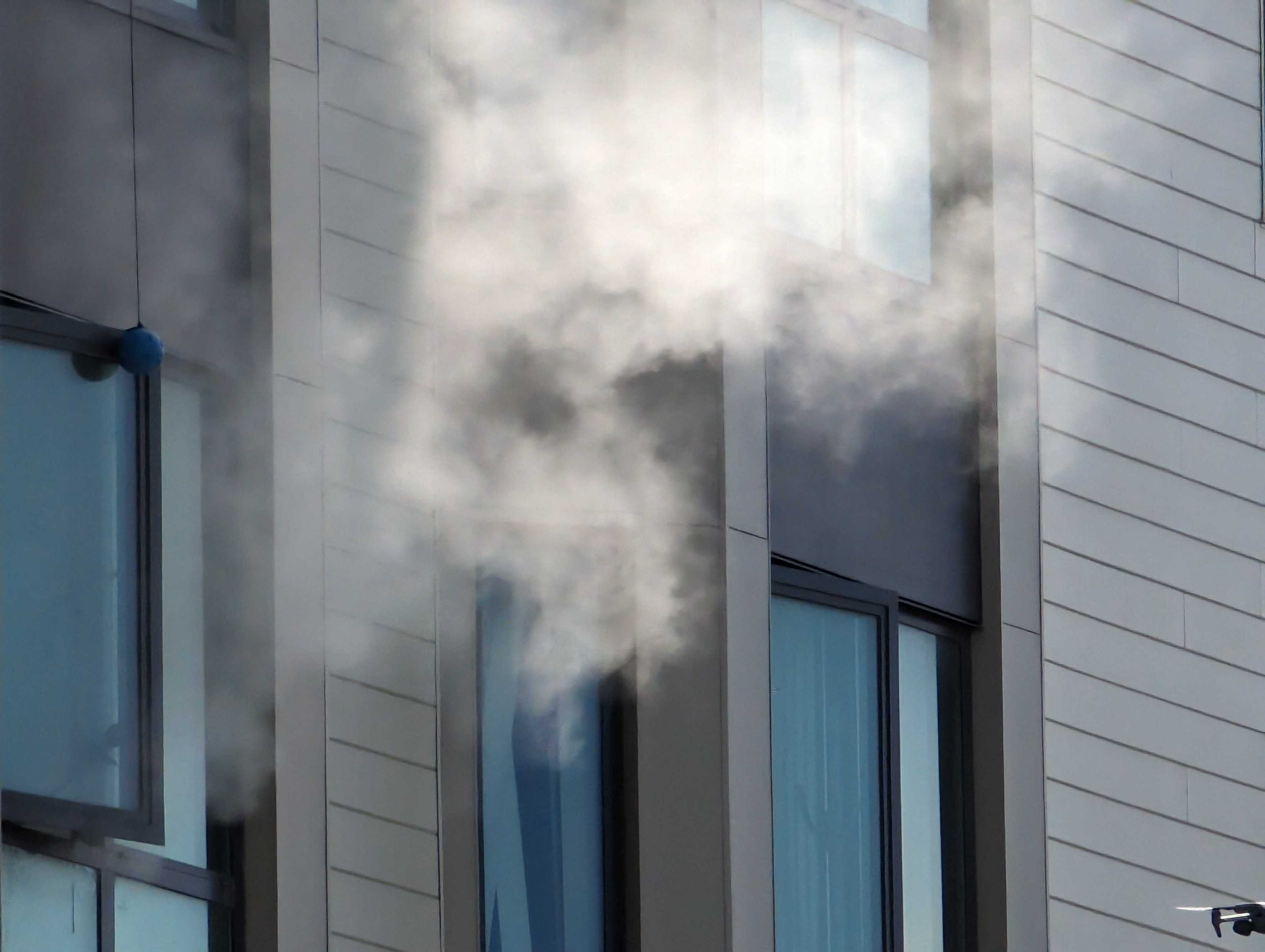 The image shows a building with light-colored siding and large windows emitting thick white smoke, suggesting a possible fire or exhaust issue.