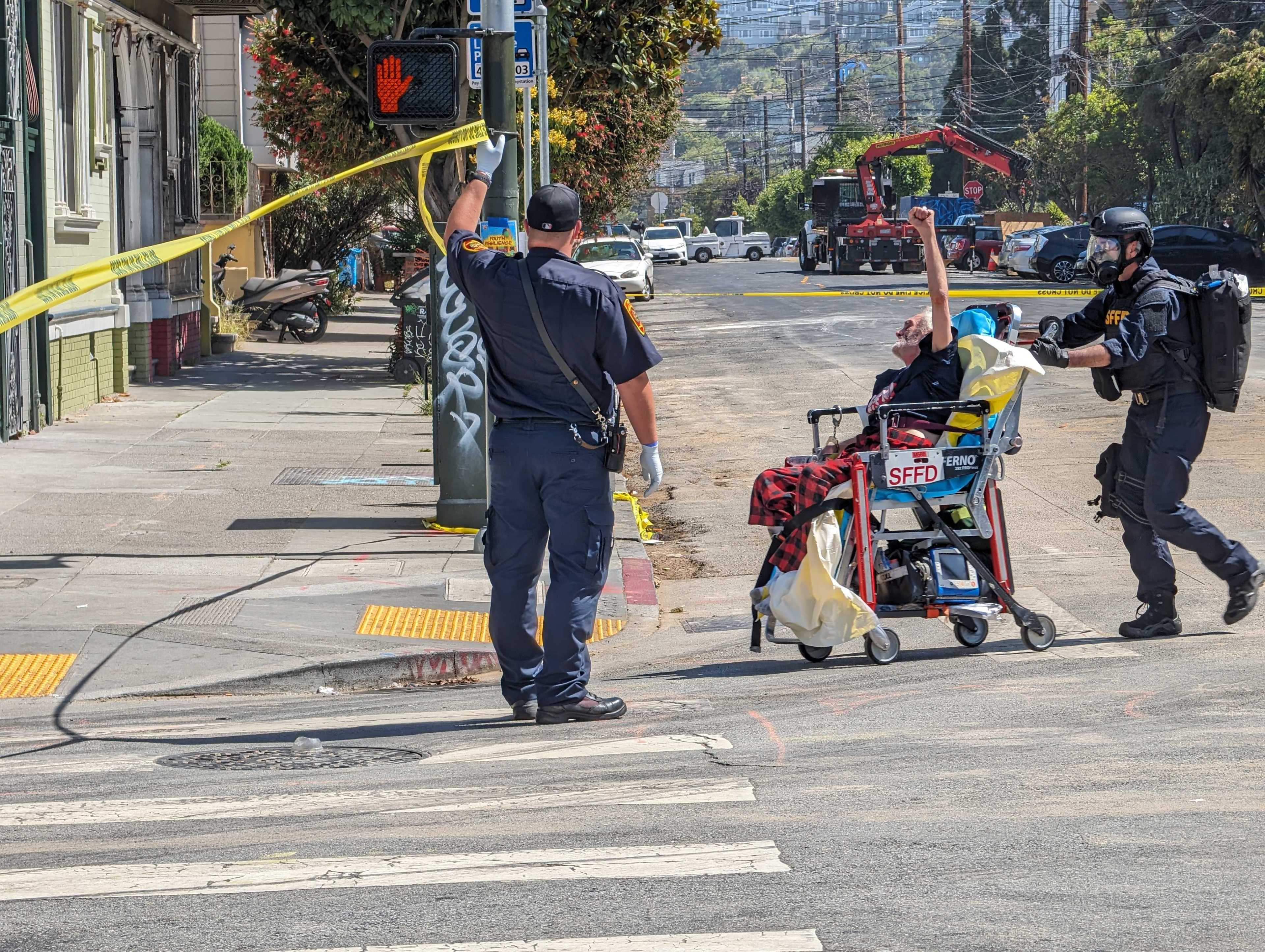 A person in a wheelchair is escorted by two officers, one holding a roll of caution tape. The street is cordoned off, with a "Do Not Walk" signal visible.