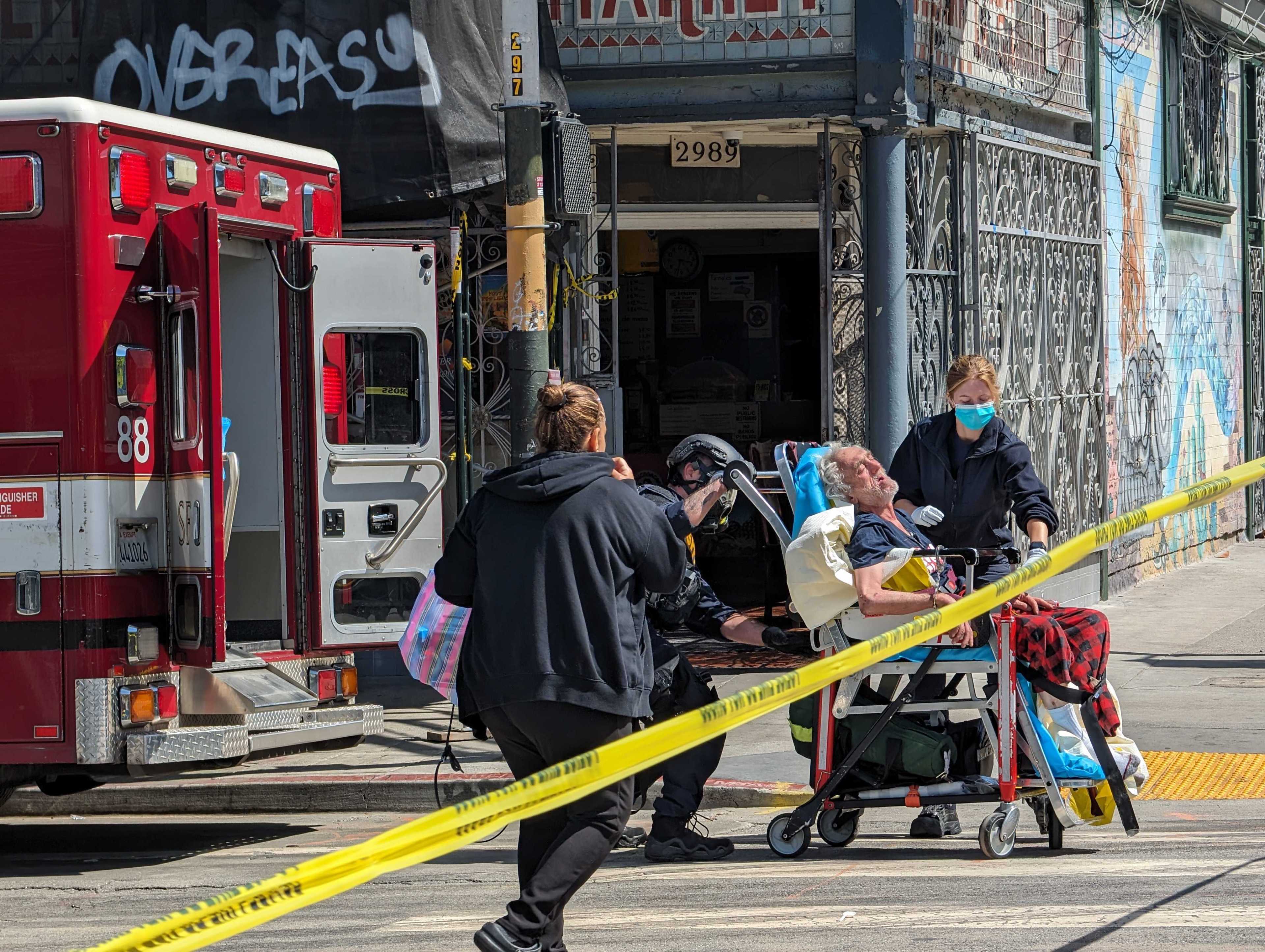 In this image, paramedics assist an elderly man on a stretcher near a red ambulance on a city street. The area is cordoned off with yellow tape, and graffiti is visible.