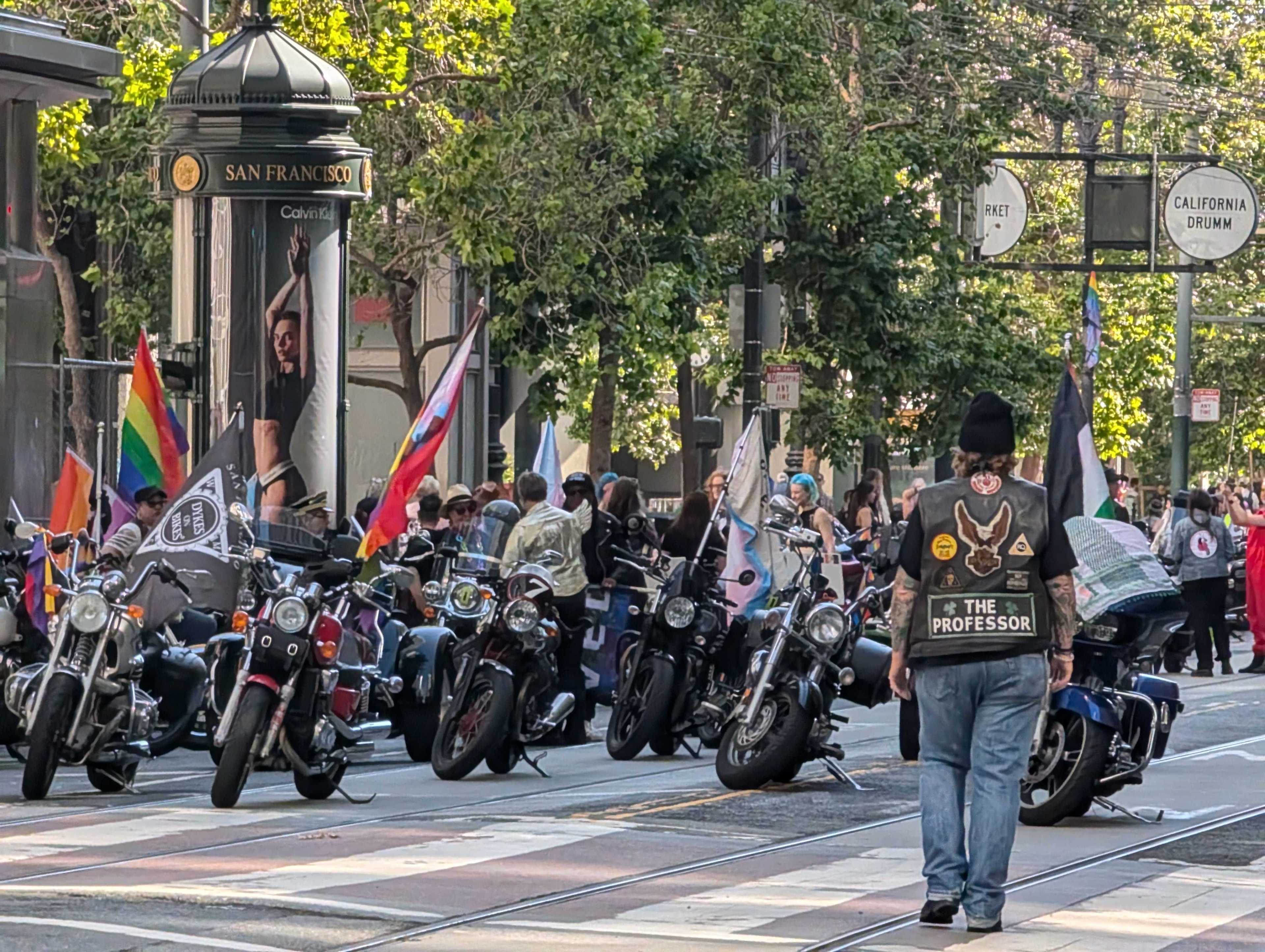 A group of motorcyclists with various flags gathers on a San Francisco street. A person with a beanie, jeans, and a patched vest reading &quot;THE PROFESSOR&quot; is in the foreground.