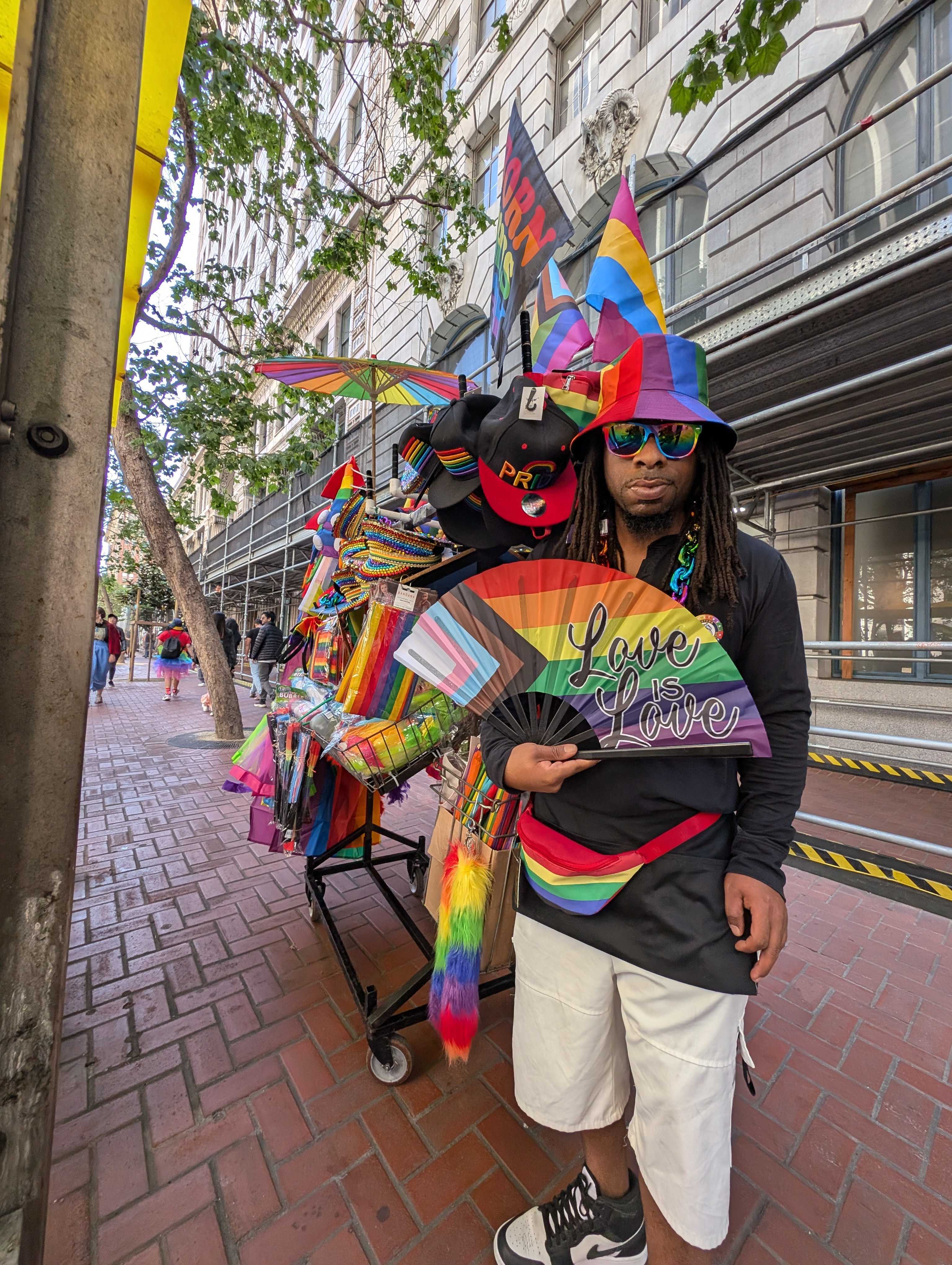 A person, dressed in colorful rainbow attire, stands by a sidewalk display filled with pride-themed items. The sign they hold reads &quot;Love is Love.&quot;