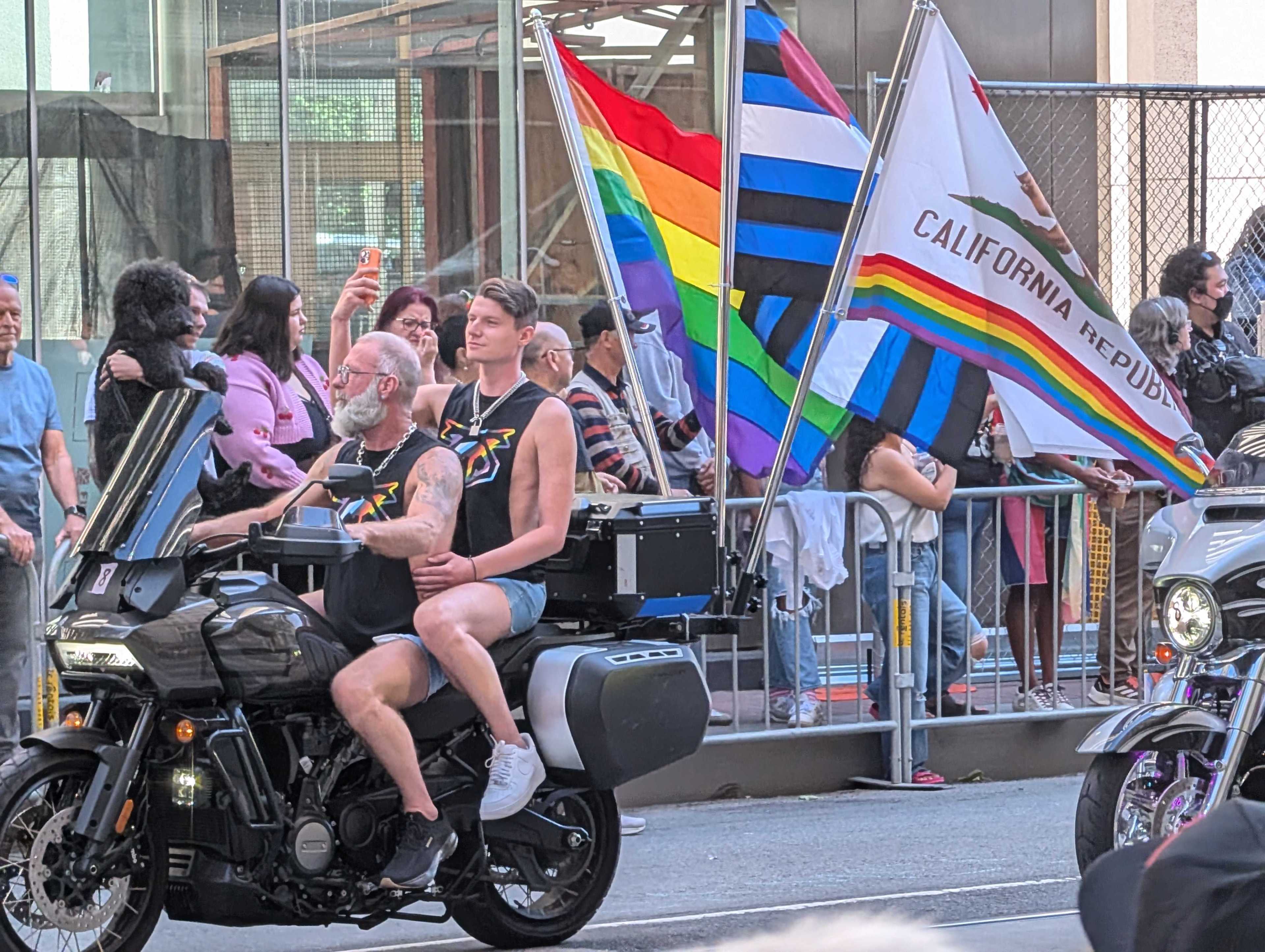Two people on a motorcycle ride in a parade, wearing matching black shirts. Behind them, a crowd holds various colorful pride flags and the California Republic flag.