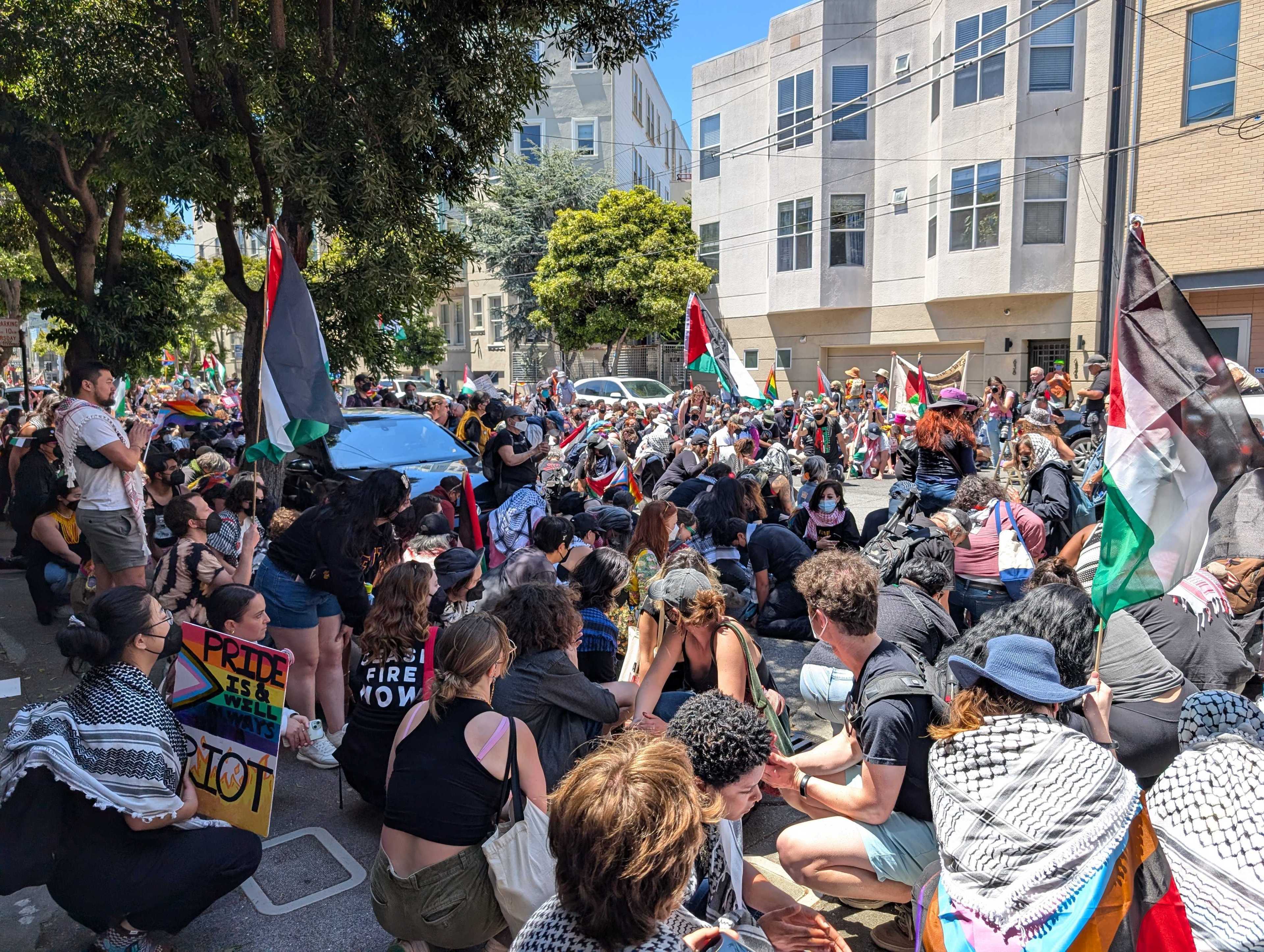 A large group of people, some holding Palestinian flags, gathers and sits on a street, with signs and banners advocating various causes. Trees and buildings are in the background.