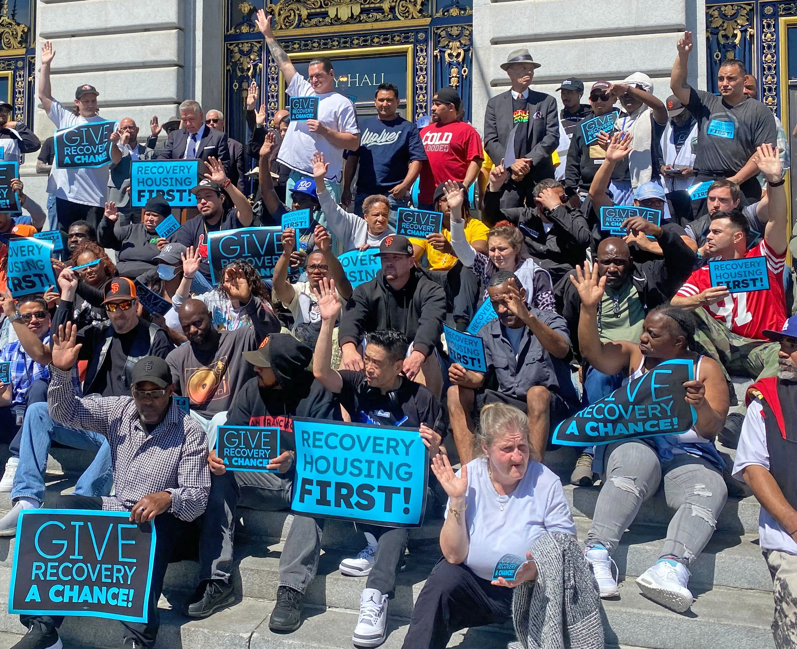 A large group of people are gathered outside, holding blue signs that read &quot;GIVE RECOVERY A CHANCE&quot; and &quot;RECOVERY HOUSING FIRST!&quot; while raising their hands.