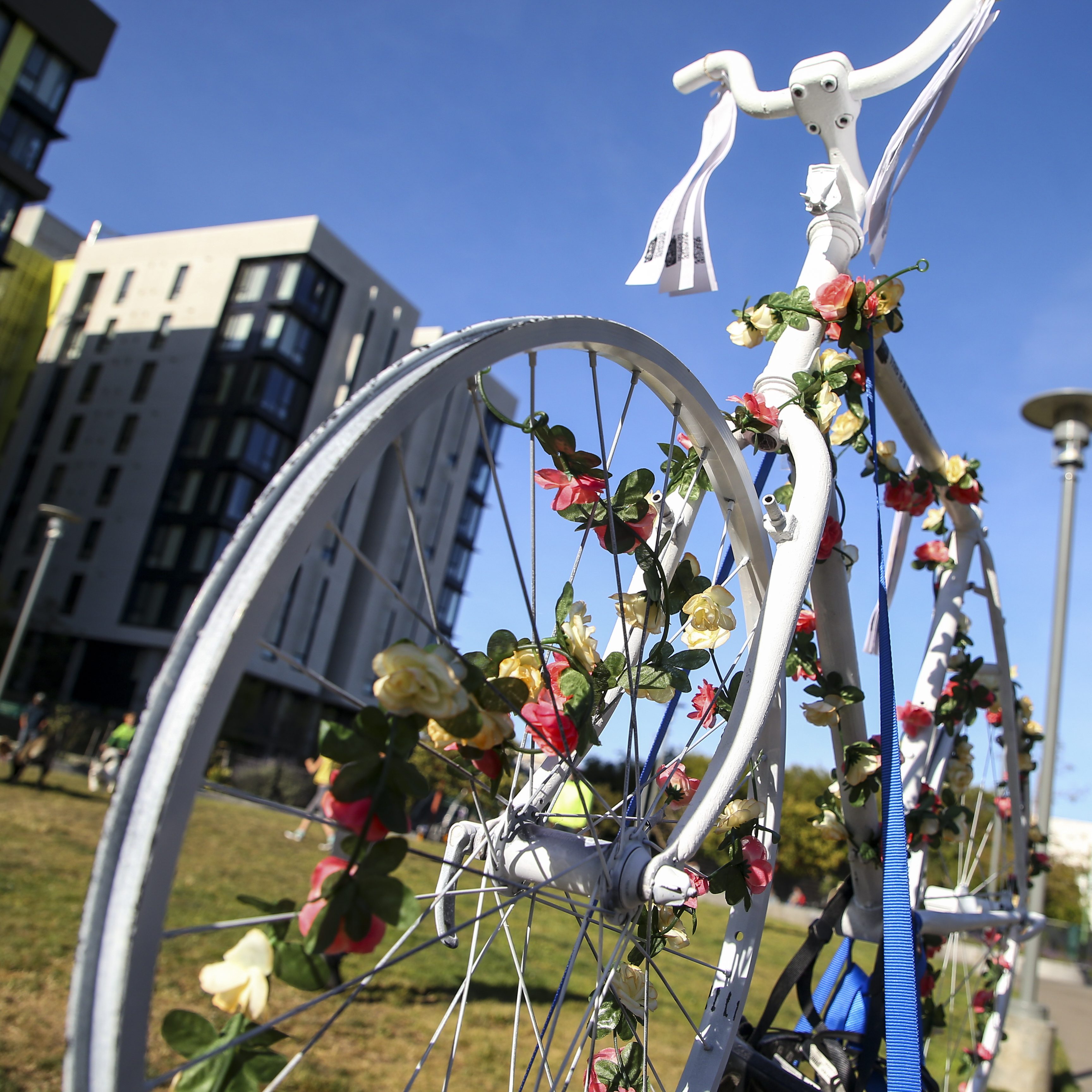 A white bicycle adorned with colorful flowers is parked in front of several modern buildings, set against a clear blue sky. A grassy area and a lamp post are also visible.