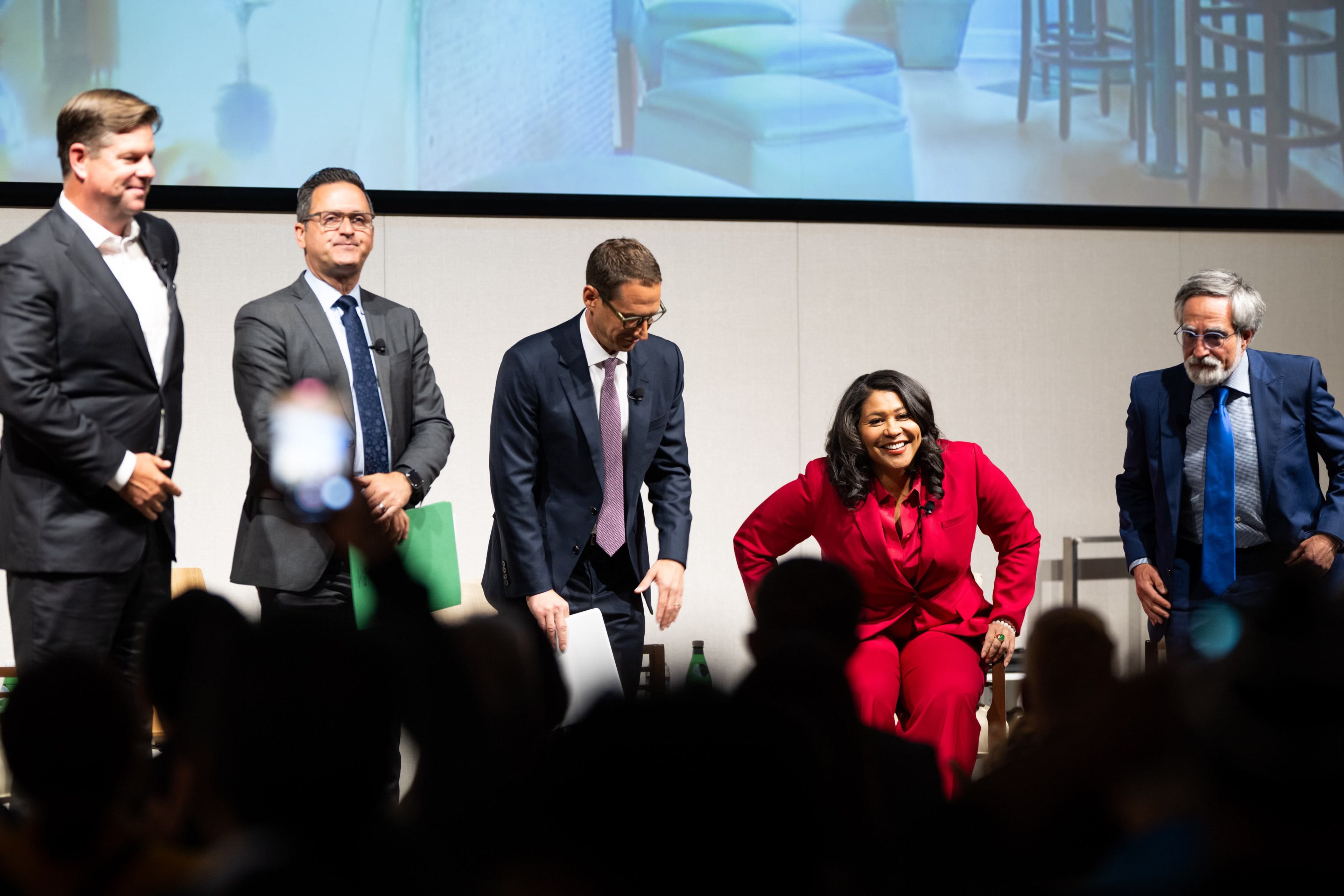 Five people, dressed formally, stand in a row on a stage in front of an audience. A woman in a red suit sits while the others remain standing.