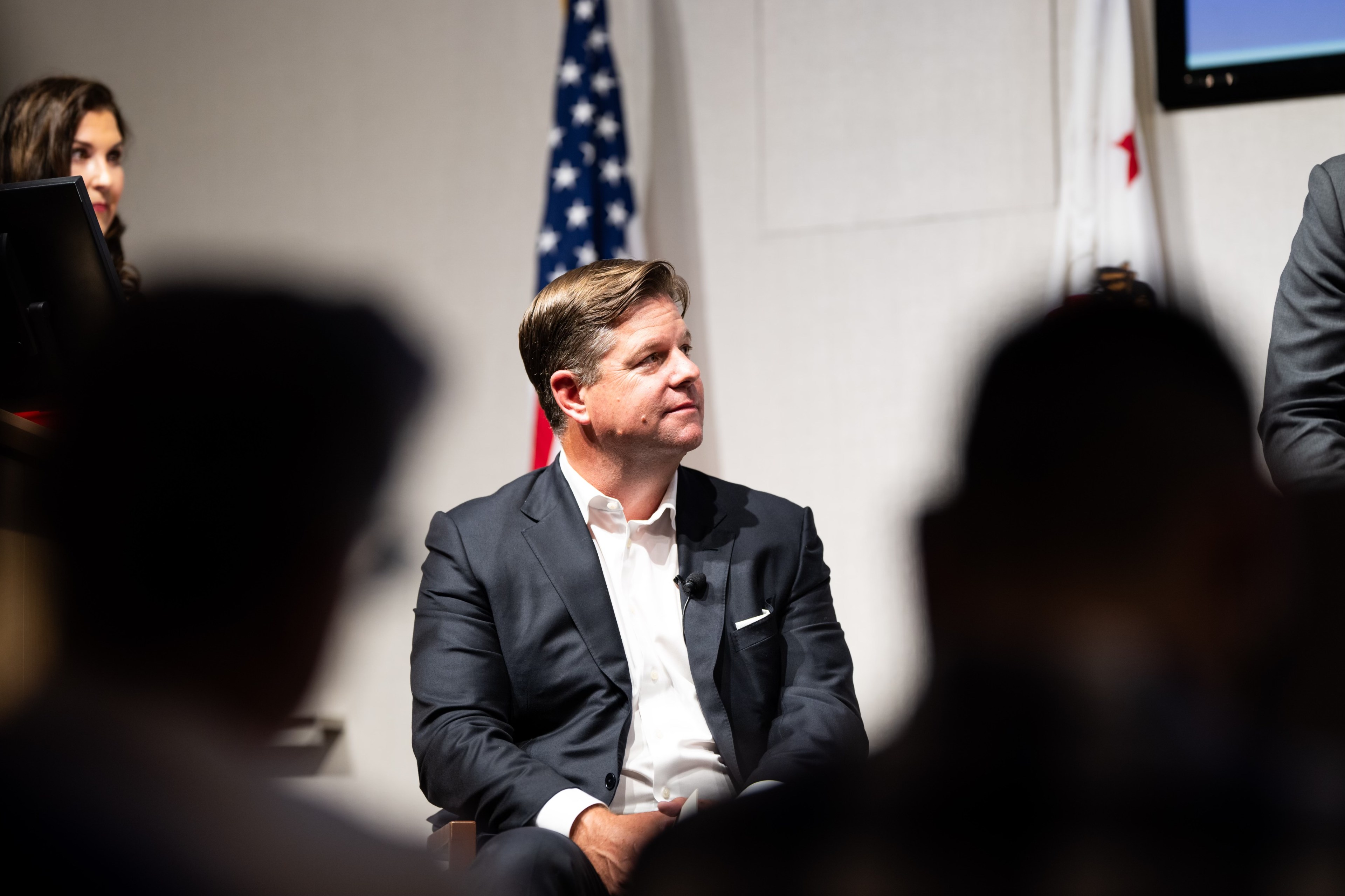 A man in a suit sits in an auditorium, facing slightly to the left, with an American flag and others in the background. The setting appears professional.