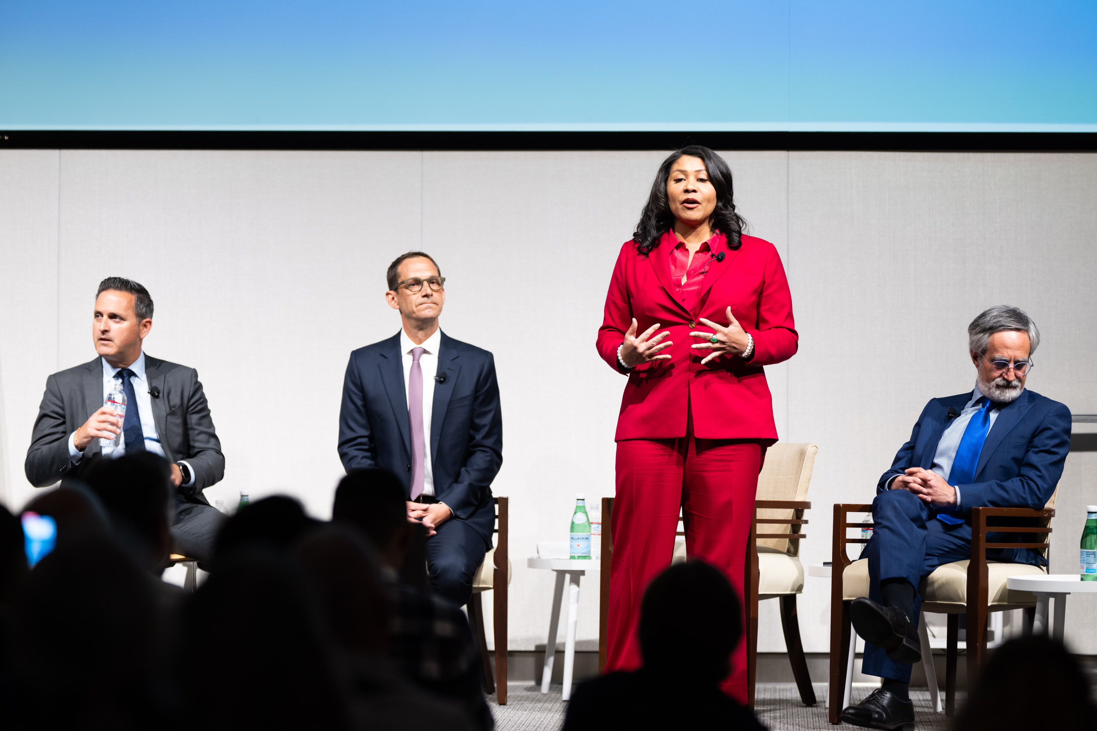 Four individuals in business attire sit on a stage; one woman in red stands talking, while three men are seated, one holding a bottle of water.