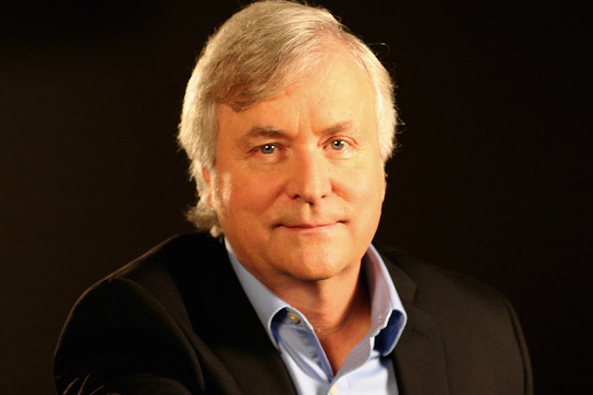 David Talbot with light gray hair wears a black blazer over a light blue collared shirt, set against a dark background. He has a calm expression and looks directly at the camera.