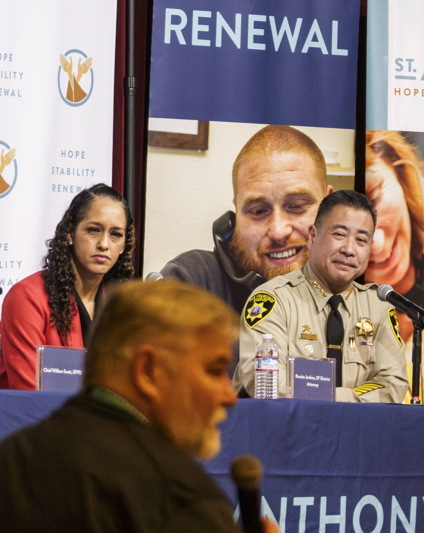 Several people sit at a table during a panel discussion. Two people are visible; one in a police uniform and another wearing a red outfit. A large backdrop displays the word &quot;RENEWAL.&quot;
