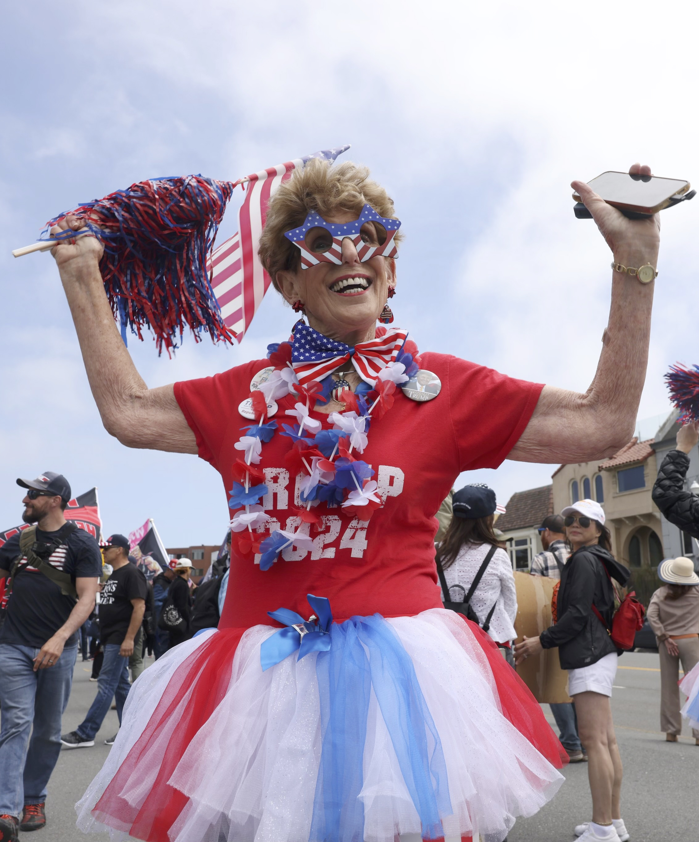 An individual in patriotic attire, featuring a red, white, and blue tutu and star-shaped glasses, is holding pom-poms and a phone during an outdoor event with other participants.