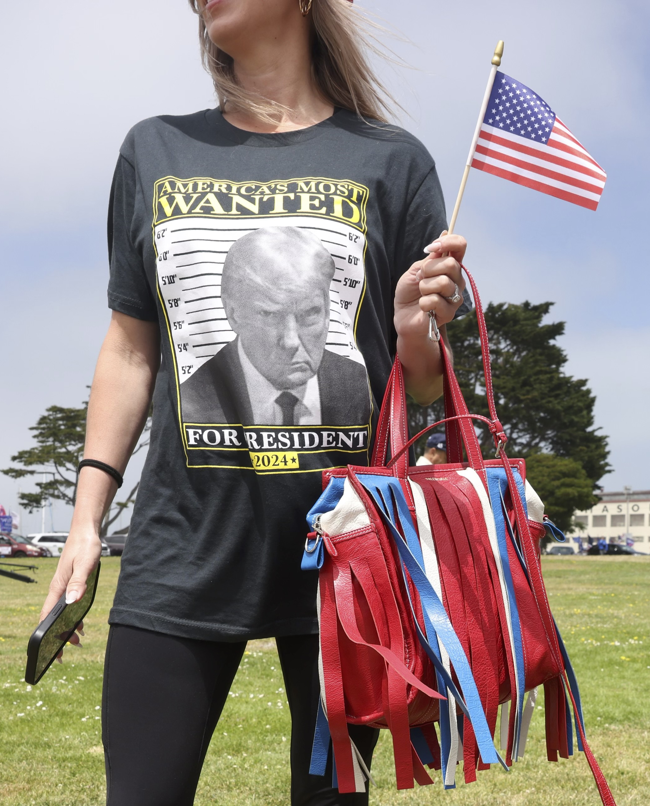 A woman wearing a black T-shirt with &quot;America's Most Wanted&quot; and an image of a man, holds a small American flag and a red fringed bag in a grassy outdoor setting.