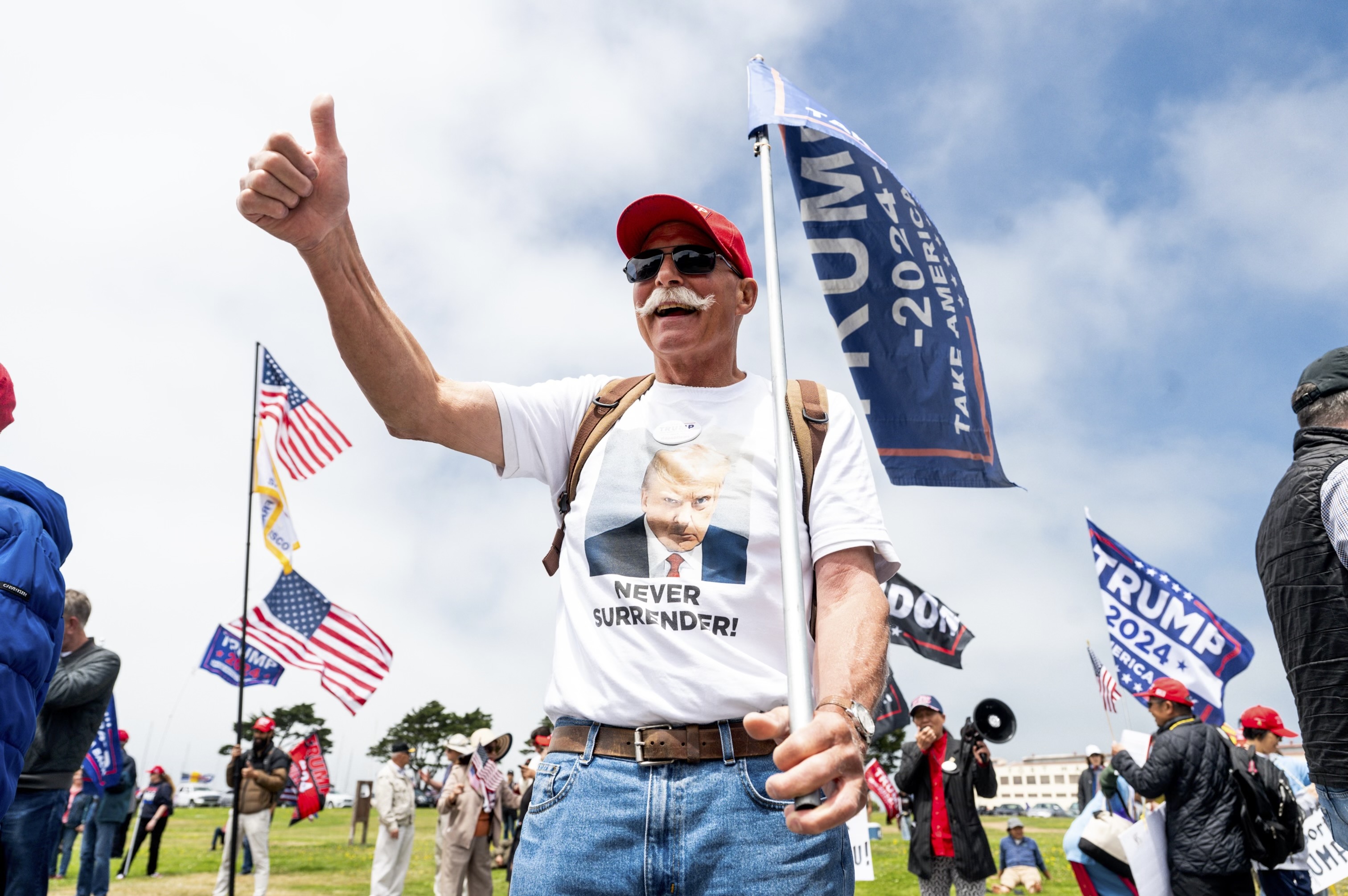 A man wearing a Trump-themed shirt and hat holds a &quot;Trump 2024&quot; flag, giving a thumbs-up in a crowd at an outdoor rally with American and Trump flags in the background.