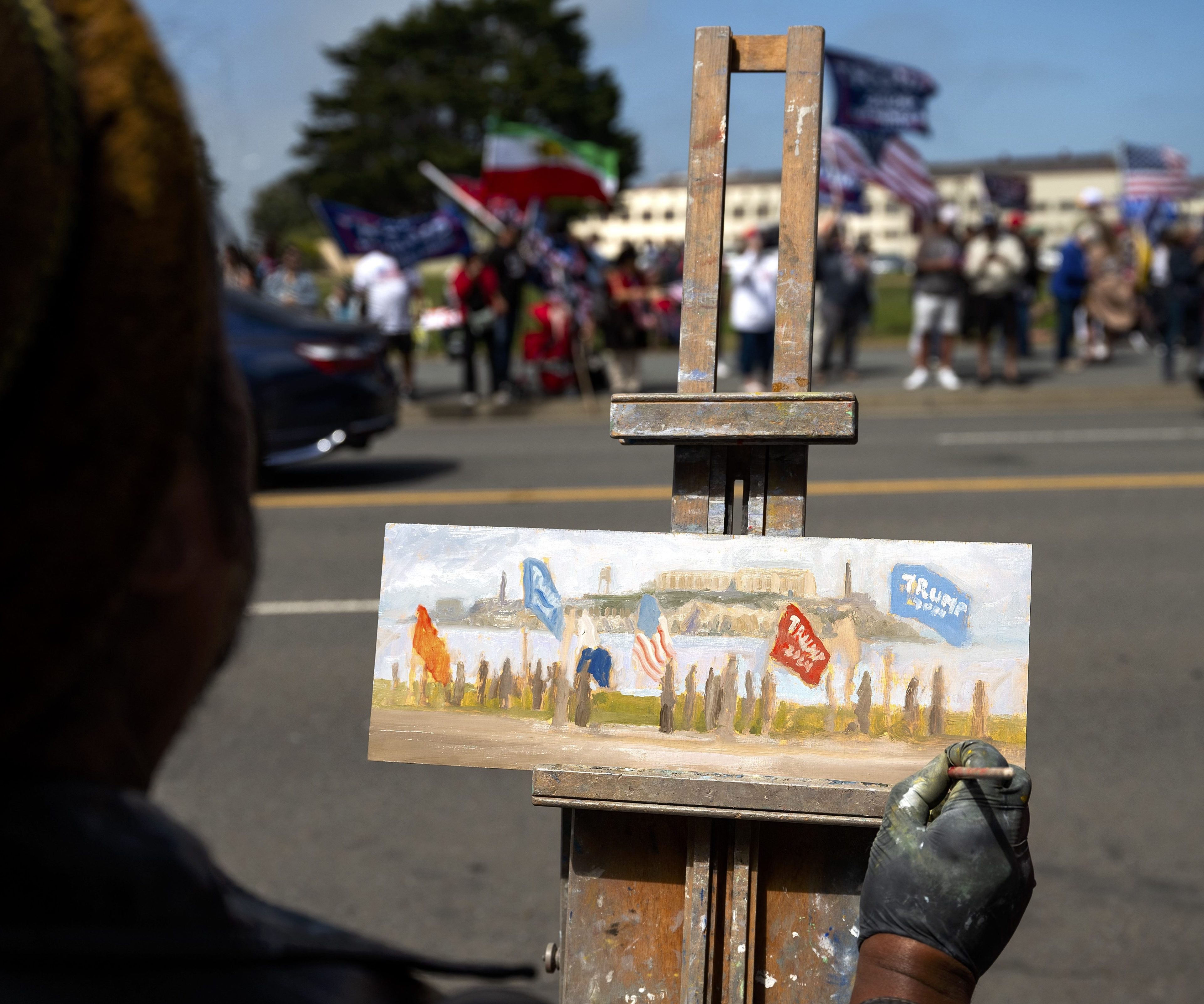 In the image, an artist paints a scene of a political rally, depicting people with flags, including &quot;Trump&quot; flags and an American flag, on a standing easel.
