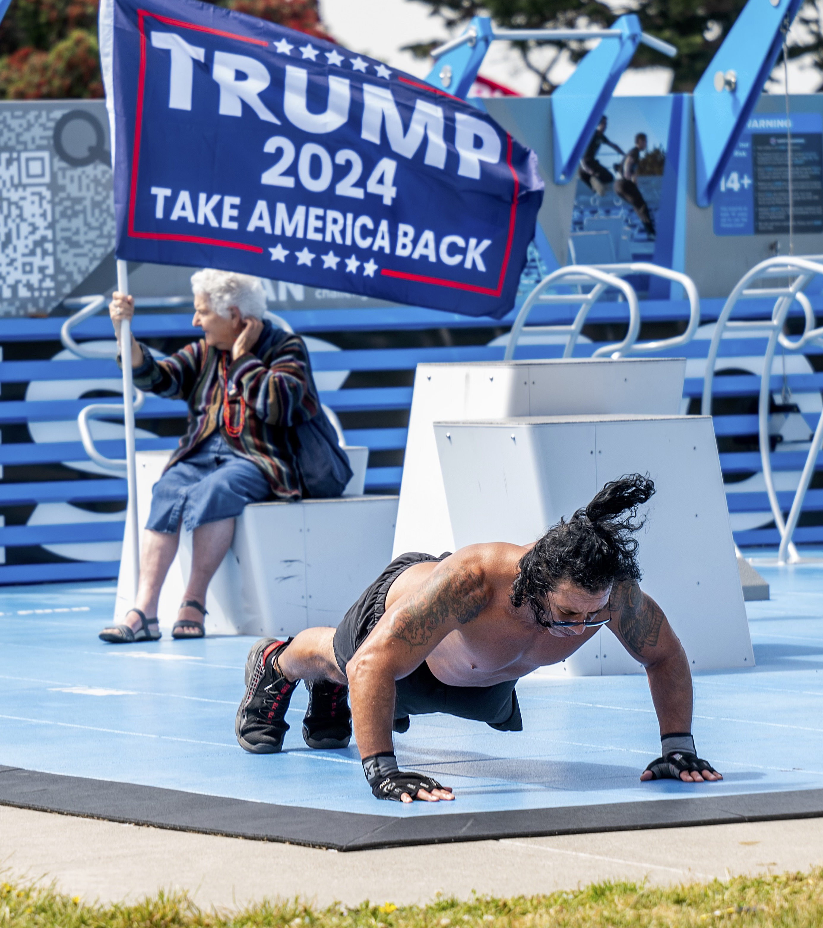 An elderly person holds a &quot;Trump 2024 Take America Back&quot; flag while sitting. In the foreground, a man is doing push-ups on a blue exercise platform.