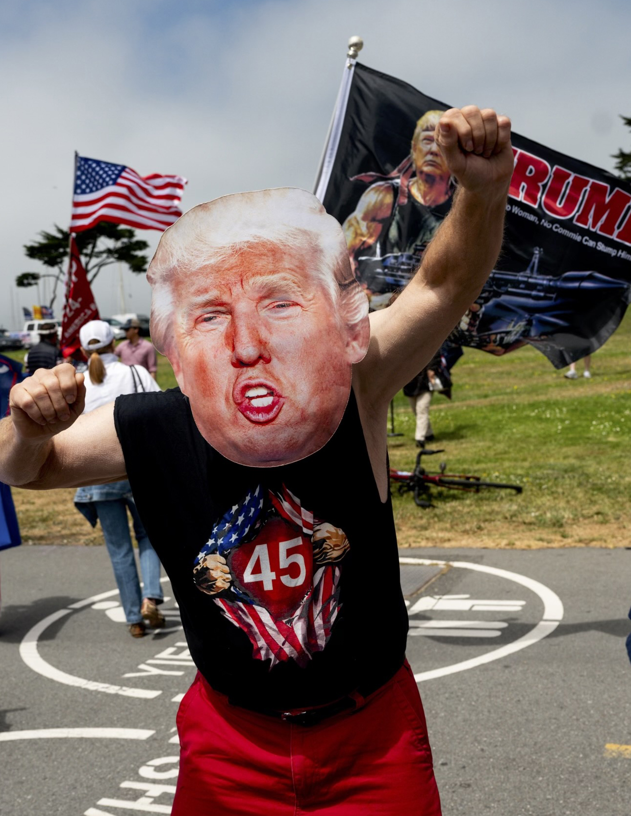 A person is wearing a mask resembling Donald Trump and a shirt depicting "45" with an American flag design. They are posing energetically, surrounded by flags in a park setting.