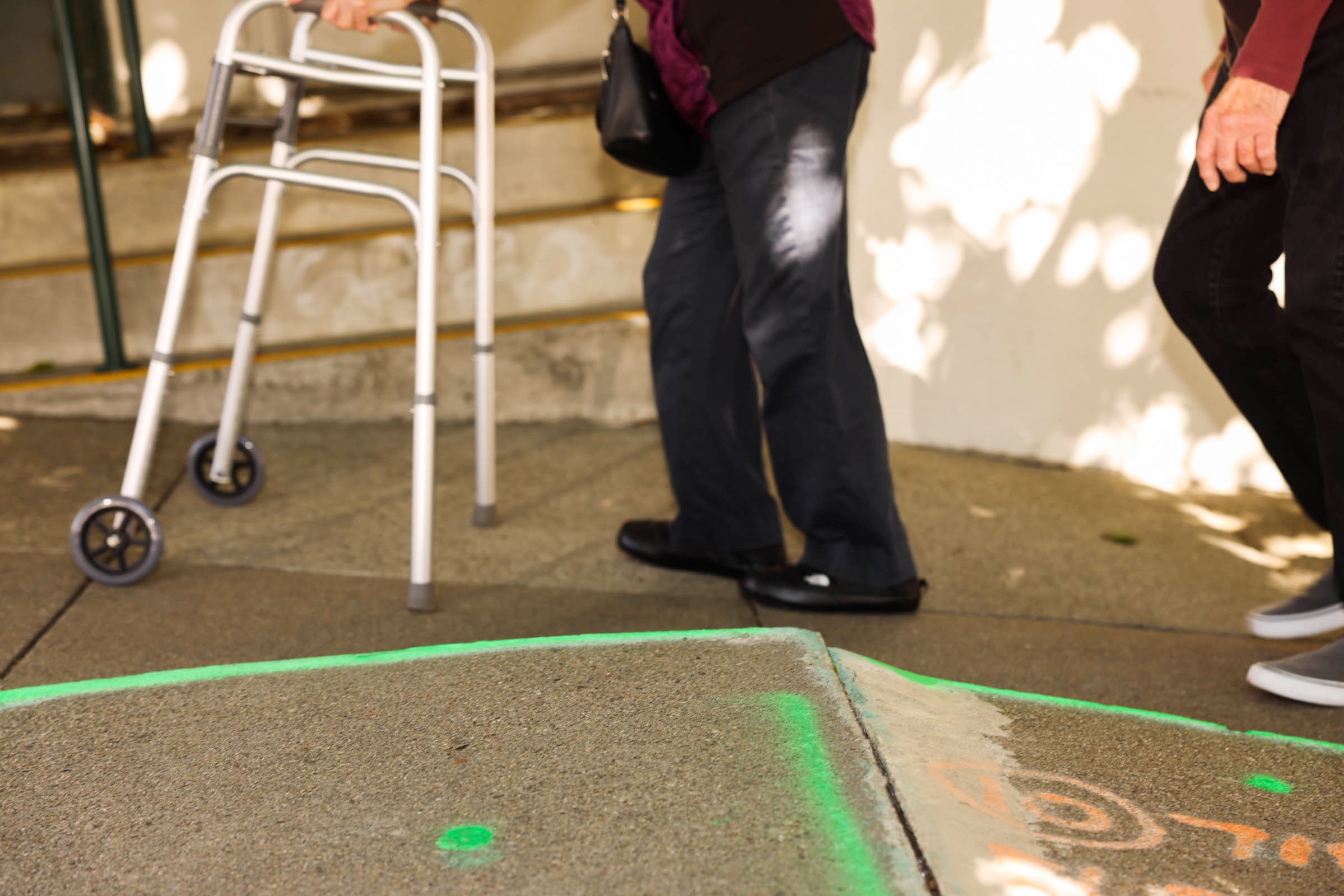 An elderly person uses a walker on a sidewalk ramp, accompanied by someone. The concrete is marked with green and orange paint, and stairs are in the background.