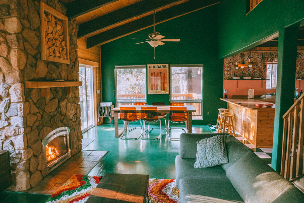 A cozy living room features a stone fireplace, green walls, a large dining table with orange chairs, and an open kitchen with wooden cabinets and bar stools.