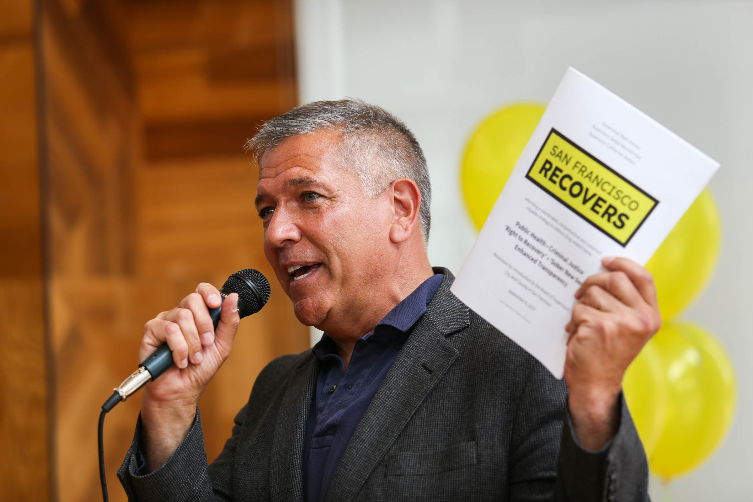 A man in a suit speaks into a microphone while holding up a document titled "San Francisco Recovers." Yellow balloons are visible in the blurred background.