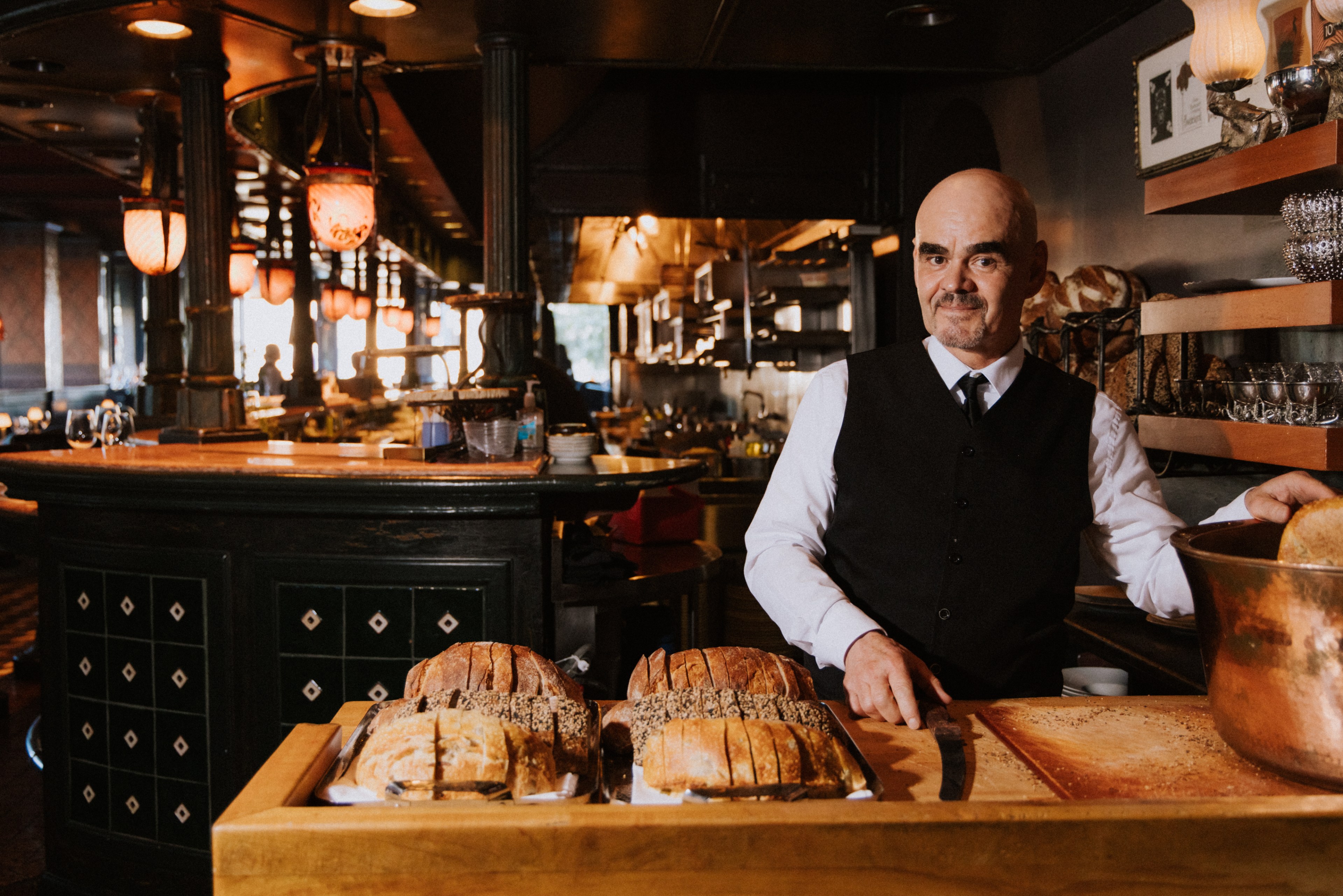 A man in a black vest and white shirt stands in a warmly lit bakery, surrounded by loaves of bread on a wooden counter, with an ornate interior in the background.