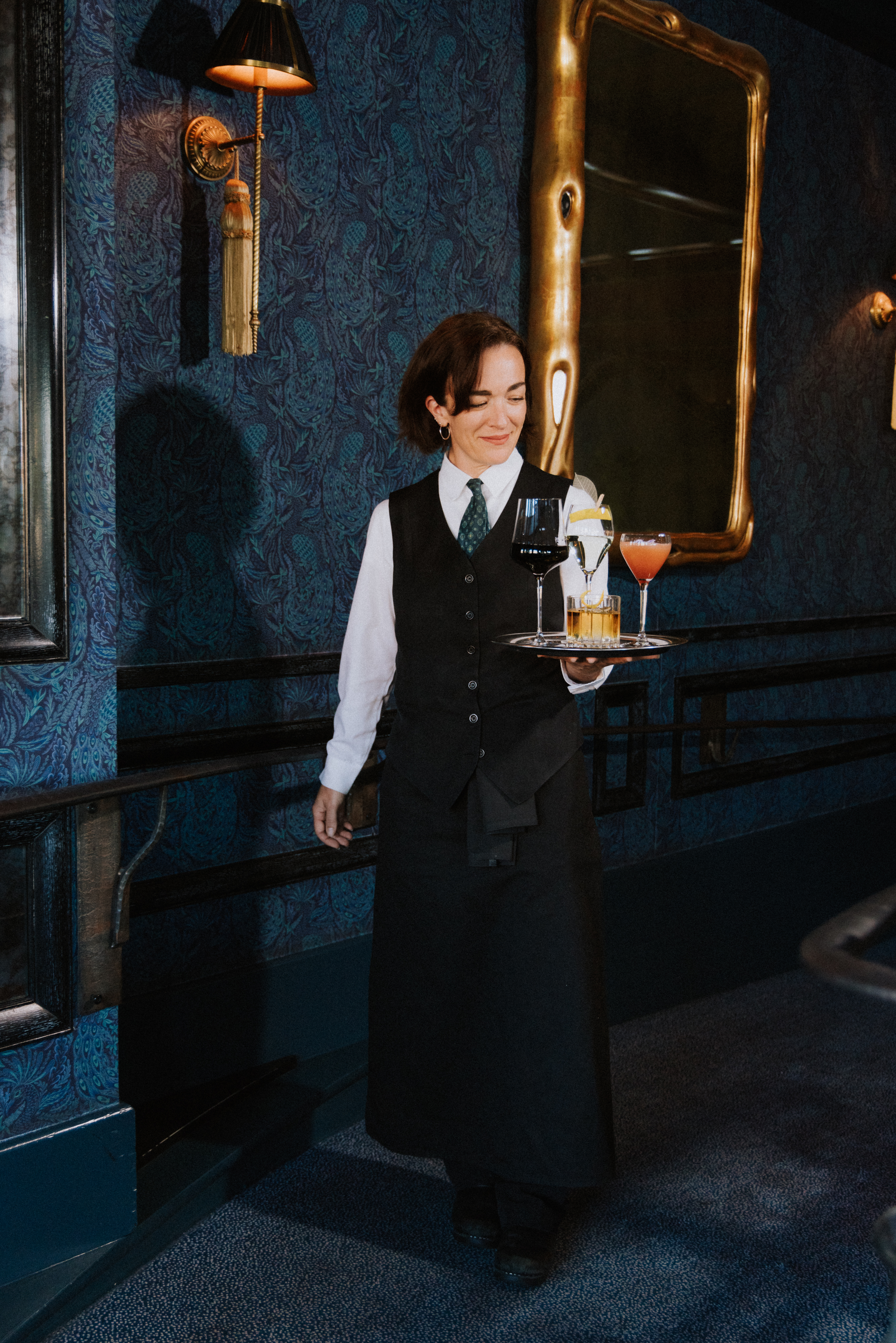 A smiling server in a black vest and tie carries a tray with four colorful drinks against an elegant, dark blue wallpaper backdrop with gold-framed mirror.