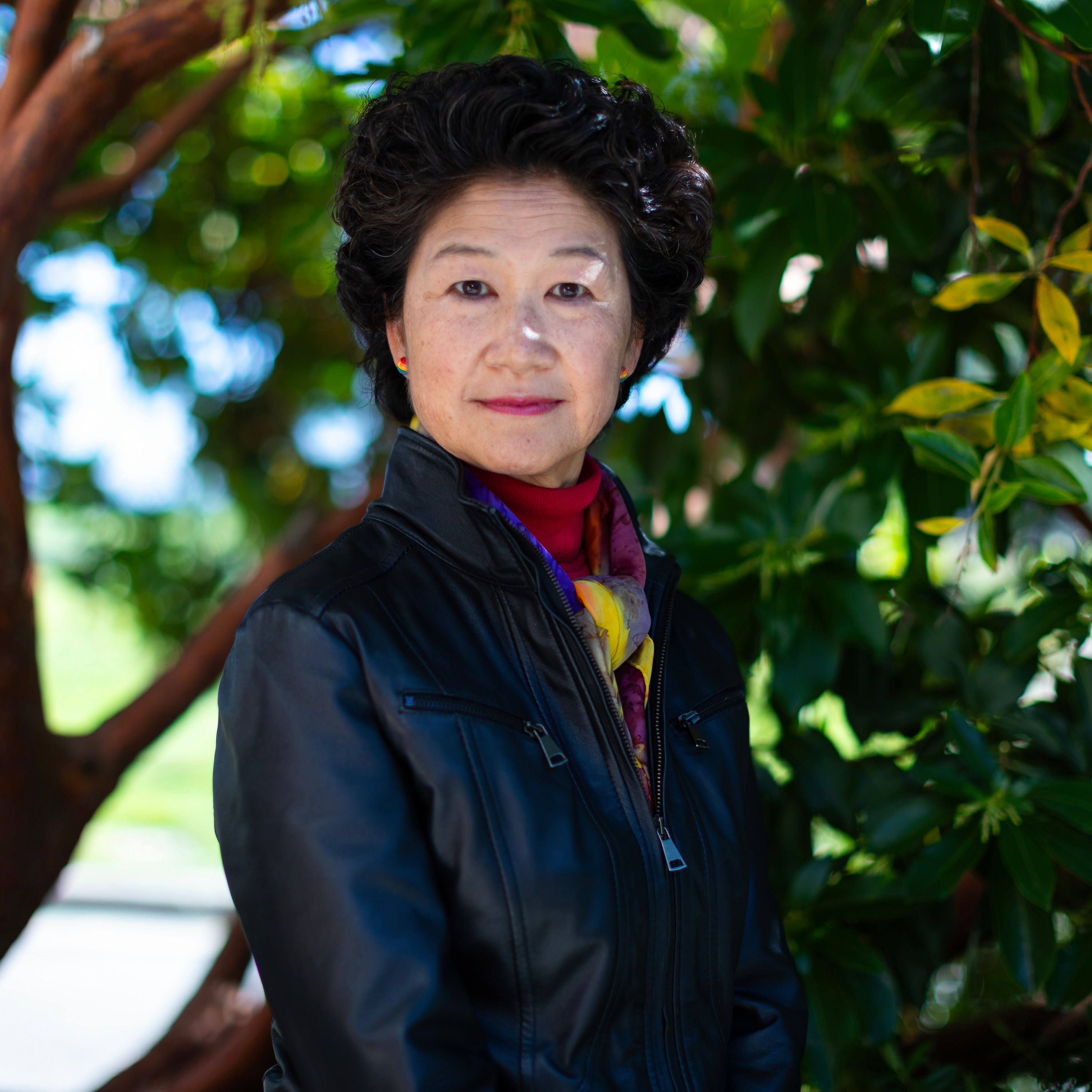 A woman in a black jacket stands outdoors near a tree with lush green leaves, looking at the camera with a neutral expression. There is sunlight filtering through the foliage.