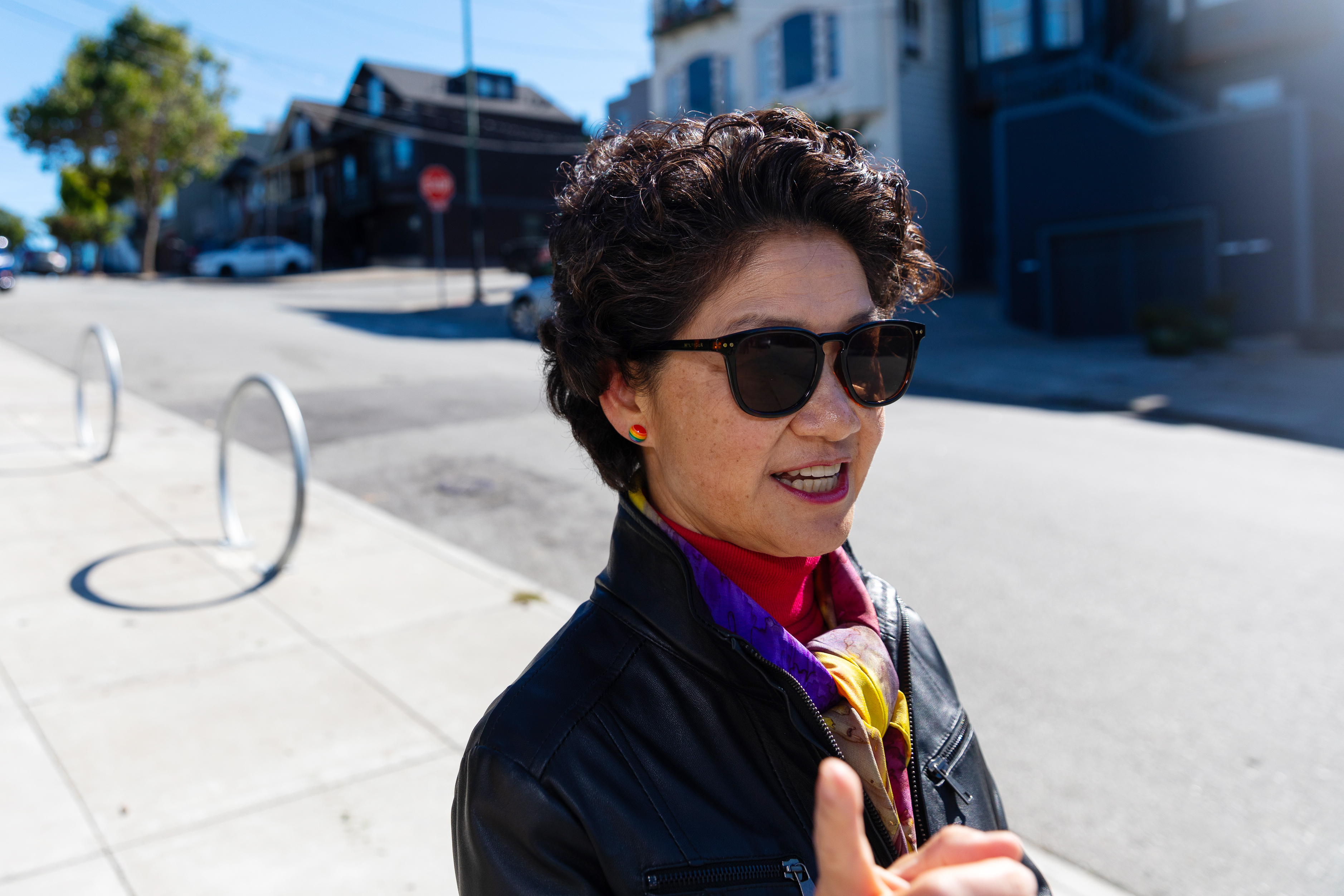 A person wearing sunglasses, a leather jacket, and a colorful scarf is standing on a sunny street with houses and a parked car in the background.