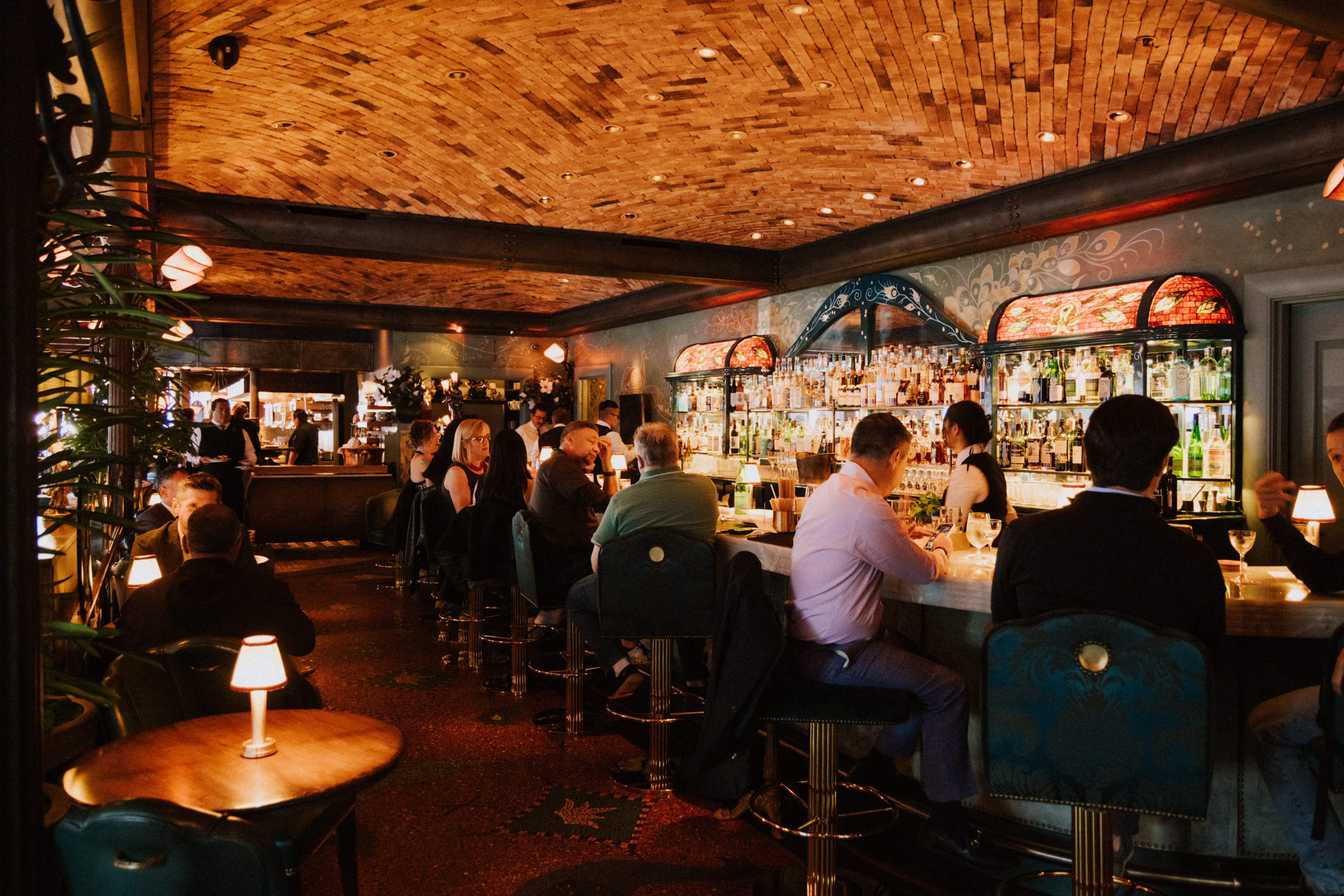 A warmly lit, bustling bar features patrons seated on barstools around a curved marble counter, with a decorative wooden ceiling and an ornate, illuminated liquor display.