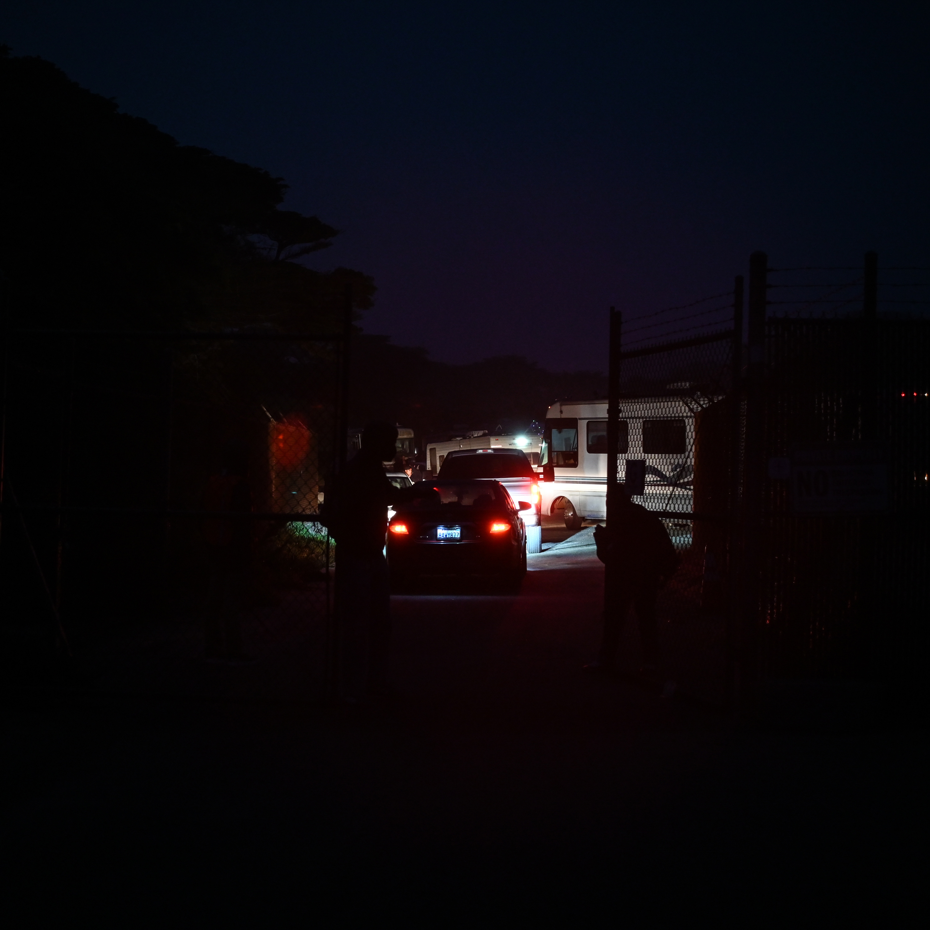 A line of vehicles drives through a gate in the dark, with headlights and taillights illuminating the scene. Silhouetted figures stand near the gate.