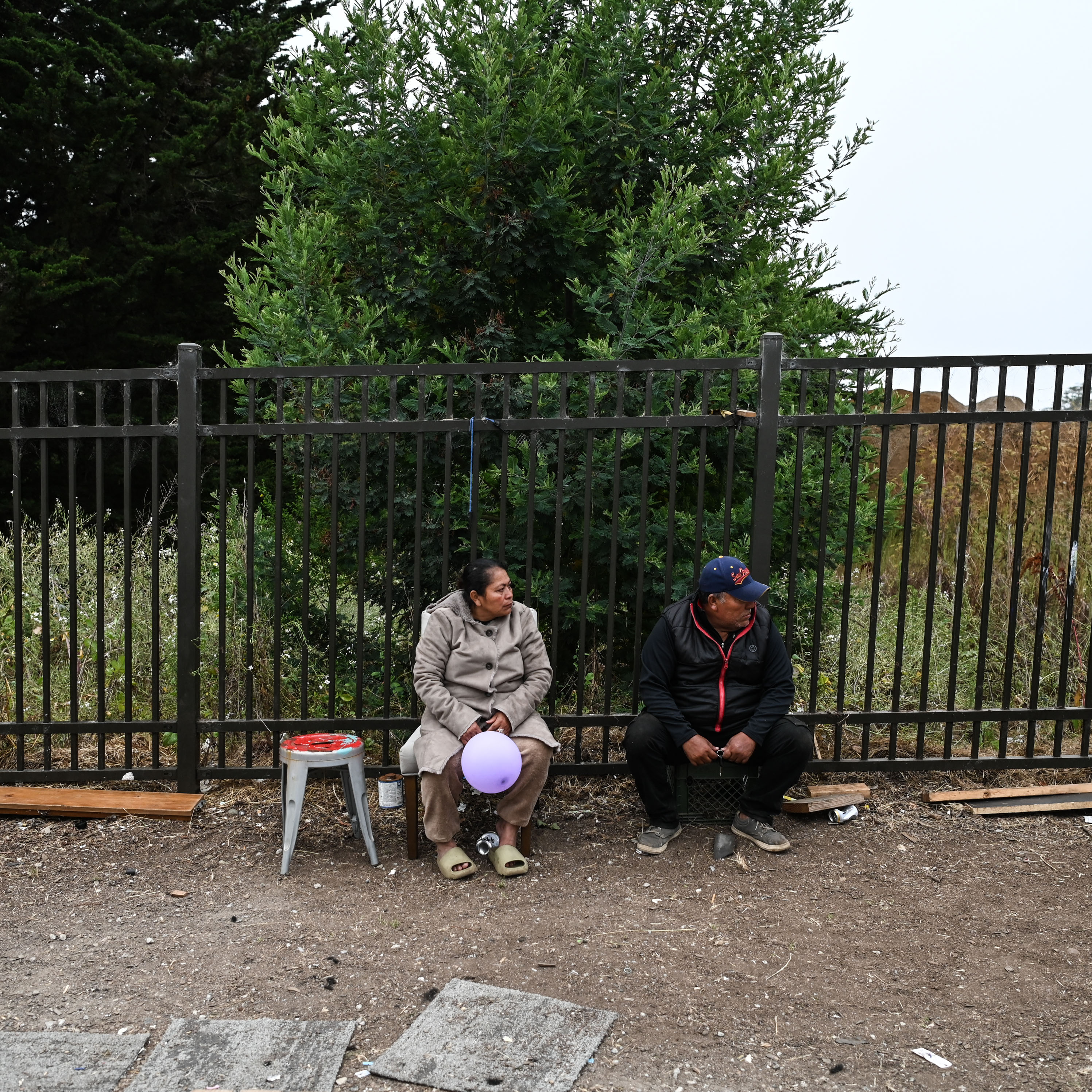 Two people sit on a ledge beside a tall metal fence, with greenery behind them. One holds a purple balloon. A small stool and a few scattered items are nearby.