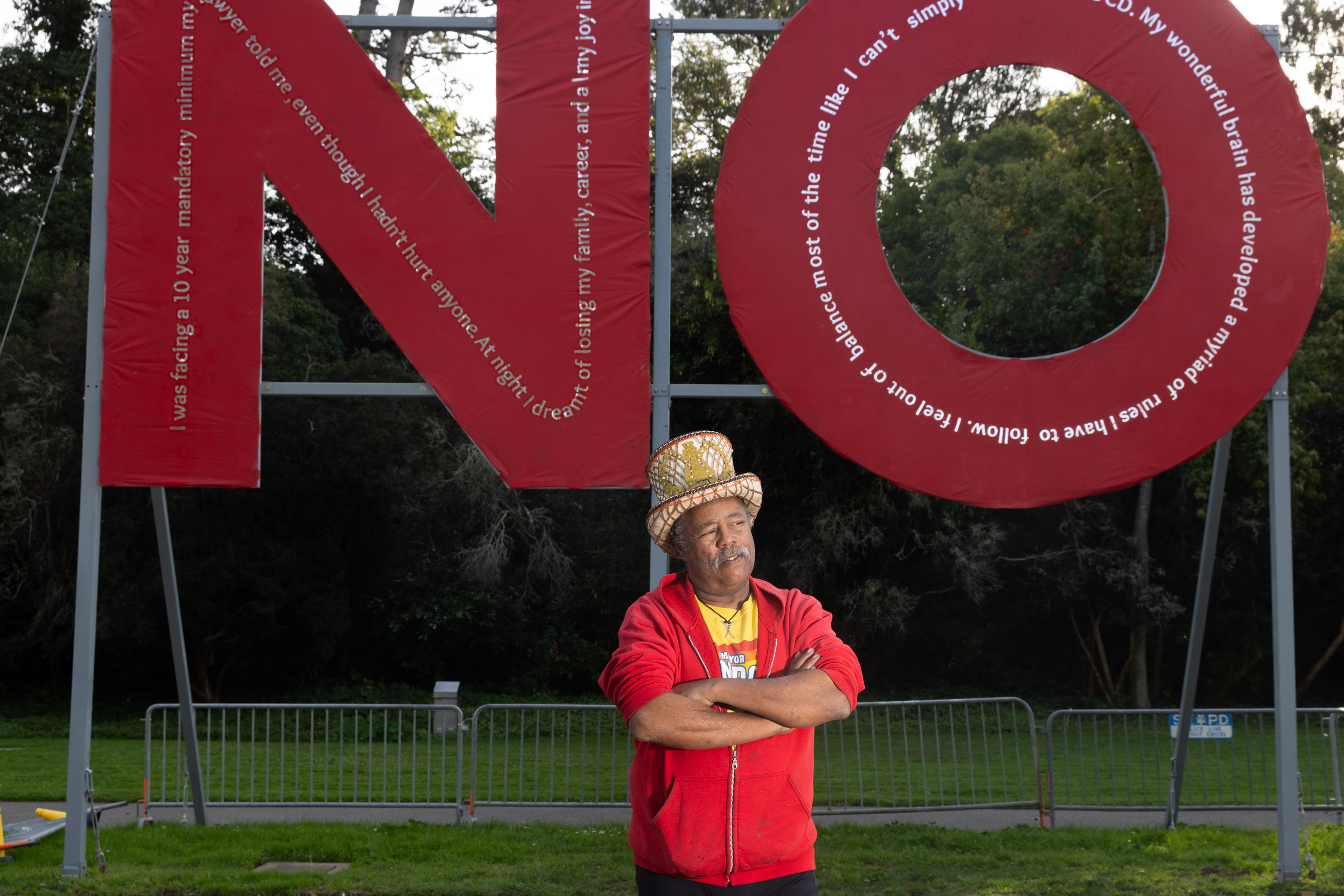 A person in a colorful hat and red hoodie stands in front of a large red structure that spells &quot;NO,&quot; with trees and metal barriers in the background.