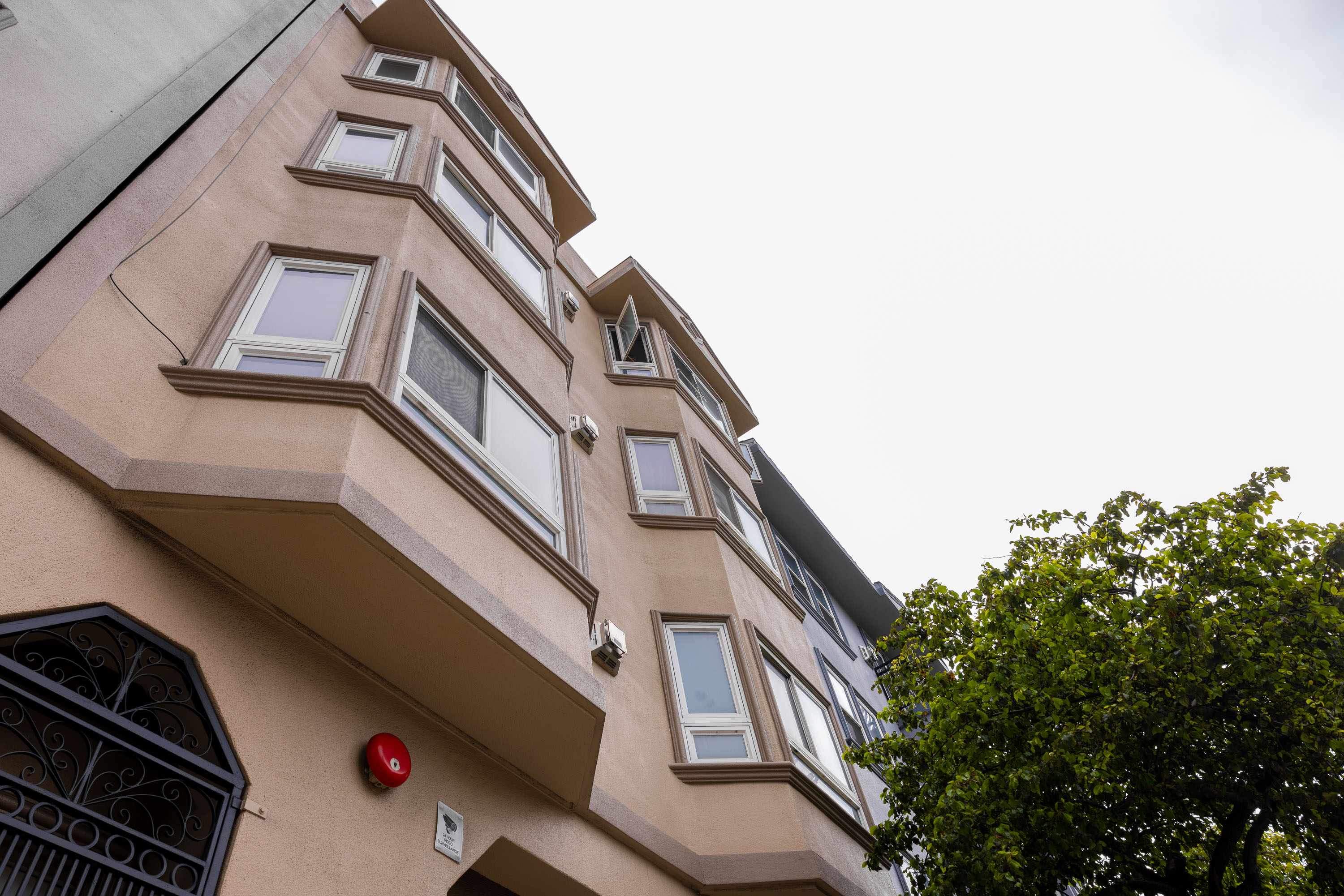 The image shows a beige multi-story building with multiple large, protruding windows. The sky is overcast, and a leafy green tree is on the right side.