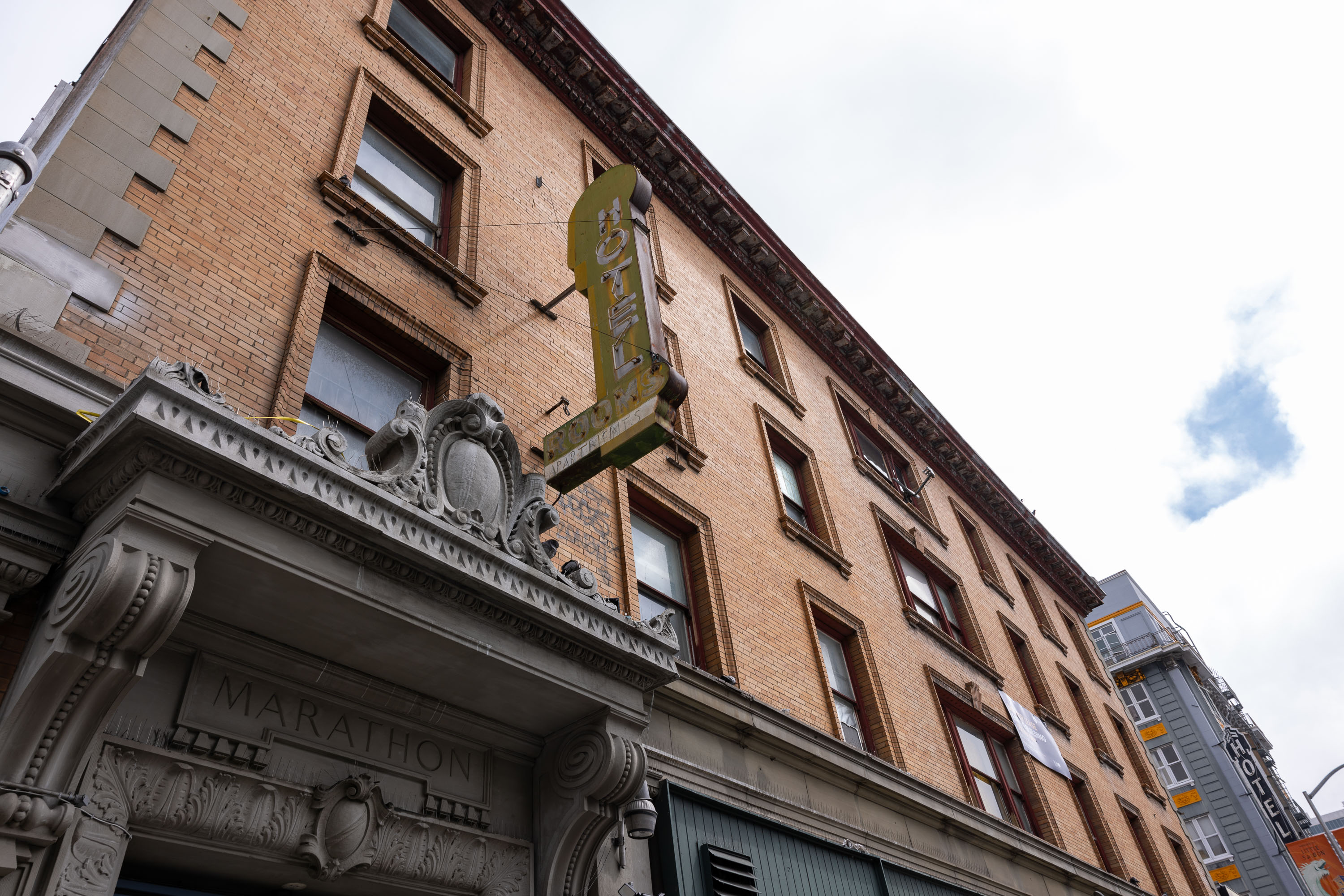 The image shows a brick building with five floors, featuring an ornate stone entrance labeled &quot;MARATHON&quot; and a vertical sign indicating &quot;HOTEL&quot; and &quot;ROOMS.&quot;