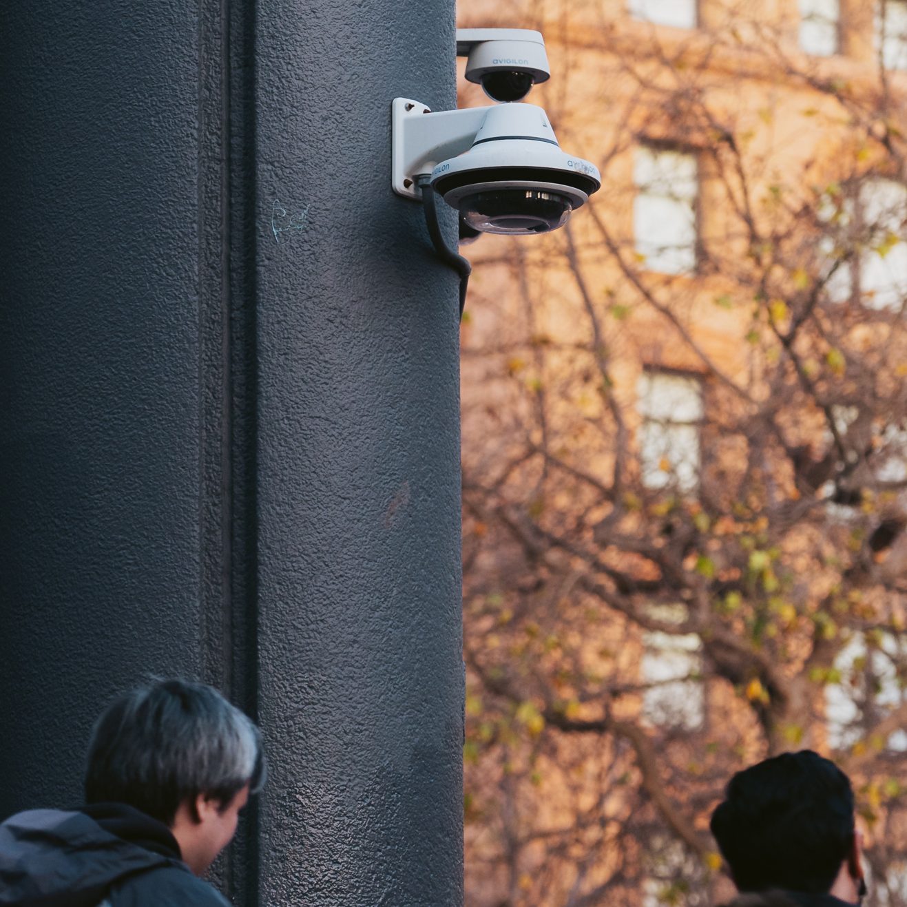 The image shows a surveillance camera mounted on a dark-colored building pillar. Two people, visible from the back, are walking past the pillar on a sunny day. 