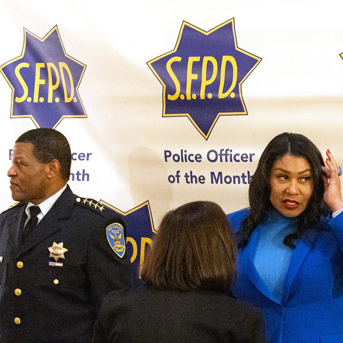 Two people stand in front of a backdrop with &quot;S.F.P.D Police Officer of the Month&quot; logos; one man in a police uniform looks left while a woman in a blue blazer looks right.