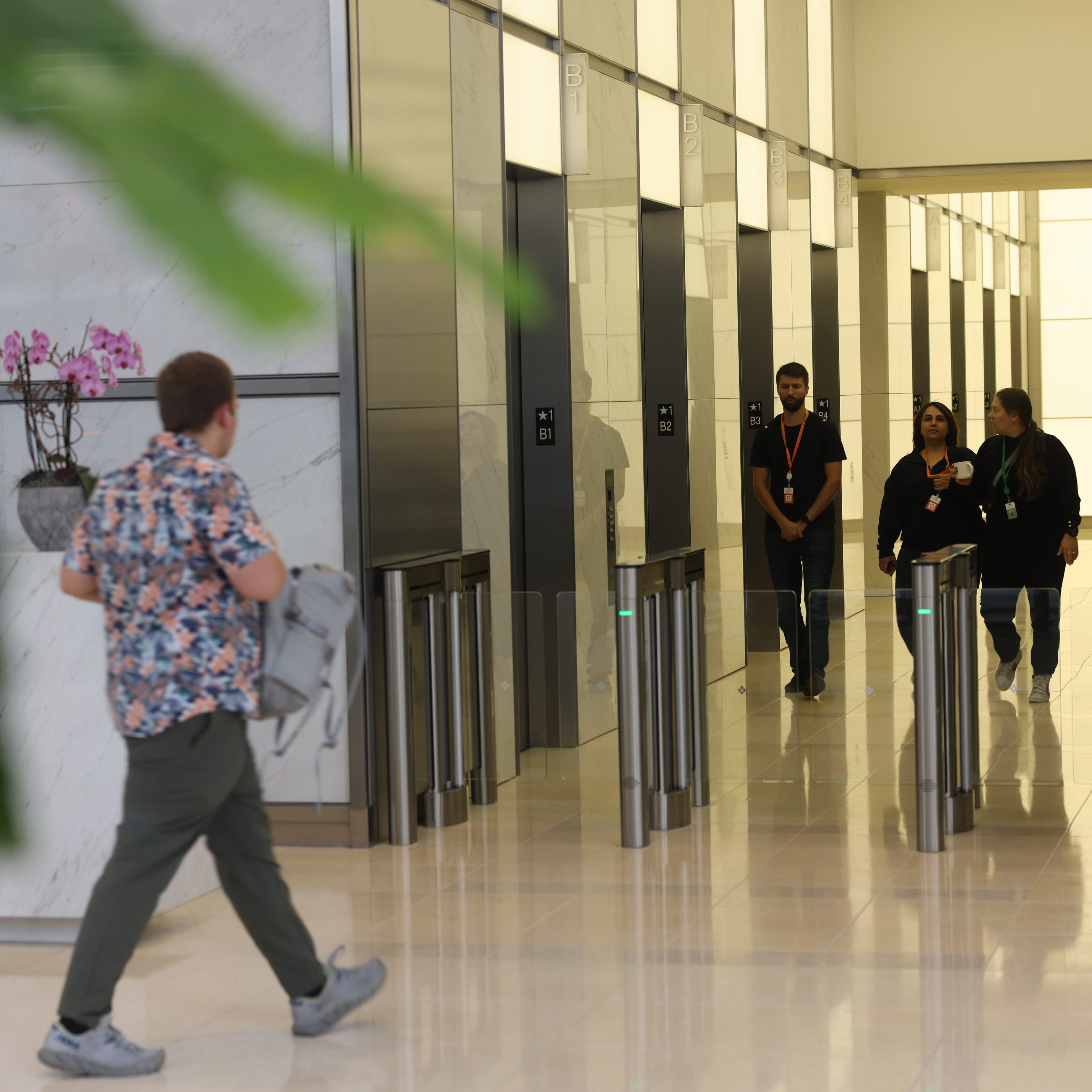 A man with a backpack walks toward a sleek elevator lobby, while two women and a man walk away from it. The lobby has modern glass turnstiles and potted orchids.