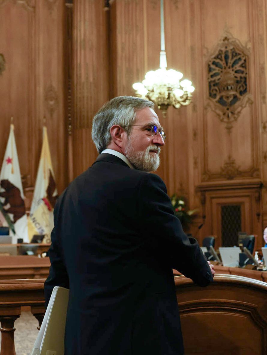 A bearded man in a suit stands at a wooden podium in an ornate room with chandeliers, flags, and seated individuals in the background, indicating a formal setting.