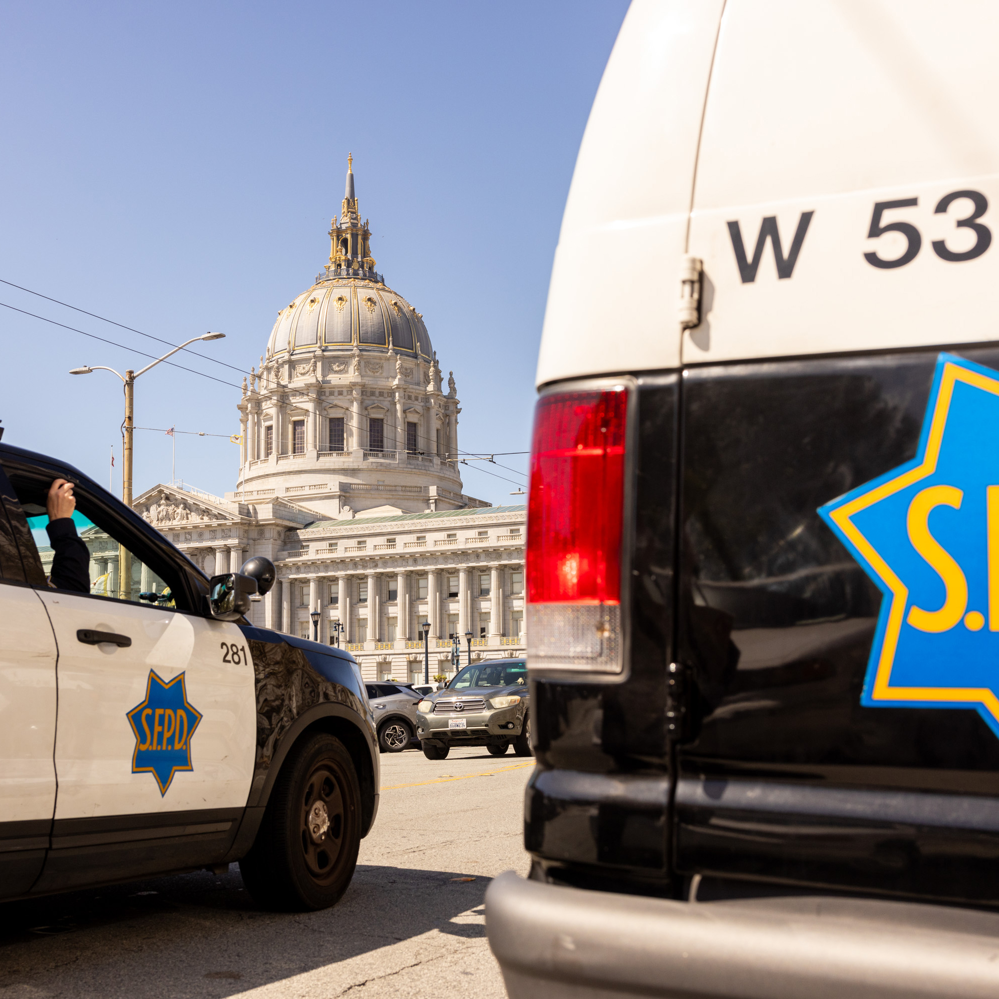 The image shows two San Francisco Police Department vehicles in the foreground, with San Francisco City Hall's domed building in the background on a sunny day.