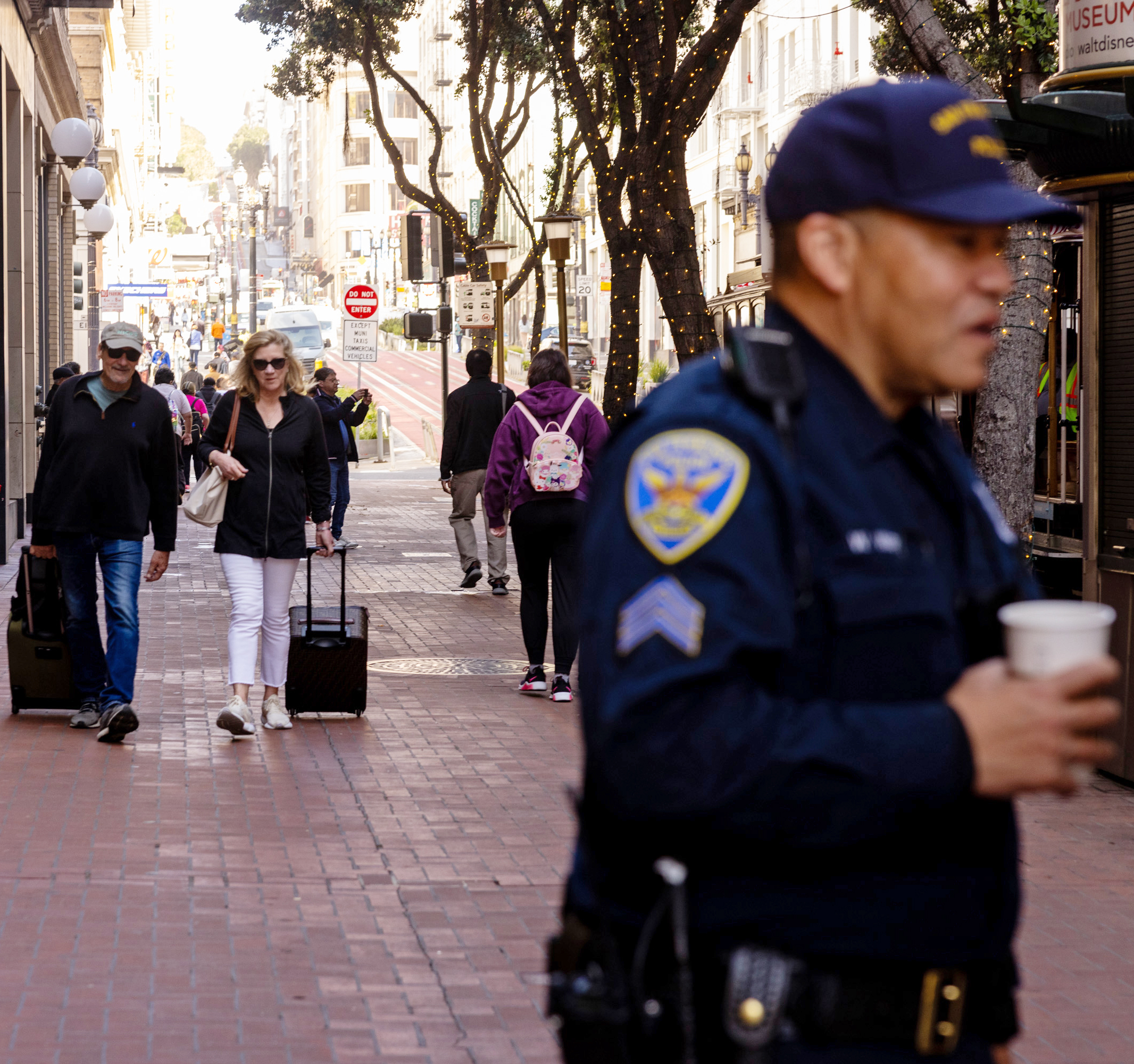 A city street with people walking, some pulling luggage. A police officer holding a cup is in the foreground, while the background shows buildings and trees with lights.